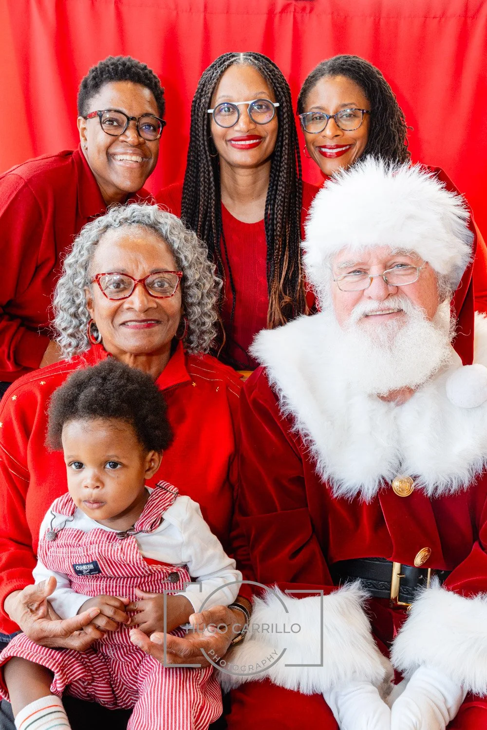 Group of six people, including Santa Claus, two women, three children, and three other women, dressed in red, posing against a red backdrop for a Christmas photo.