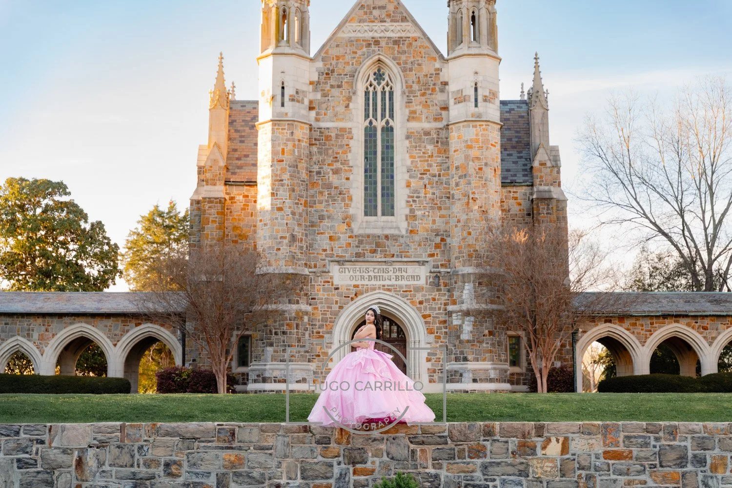 Woman in pink dress standing on a stone wall in front of a large stone church with gothic-style architecture and stained glass windows, during sunset. Trees and greenery are visible around the church.