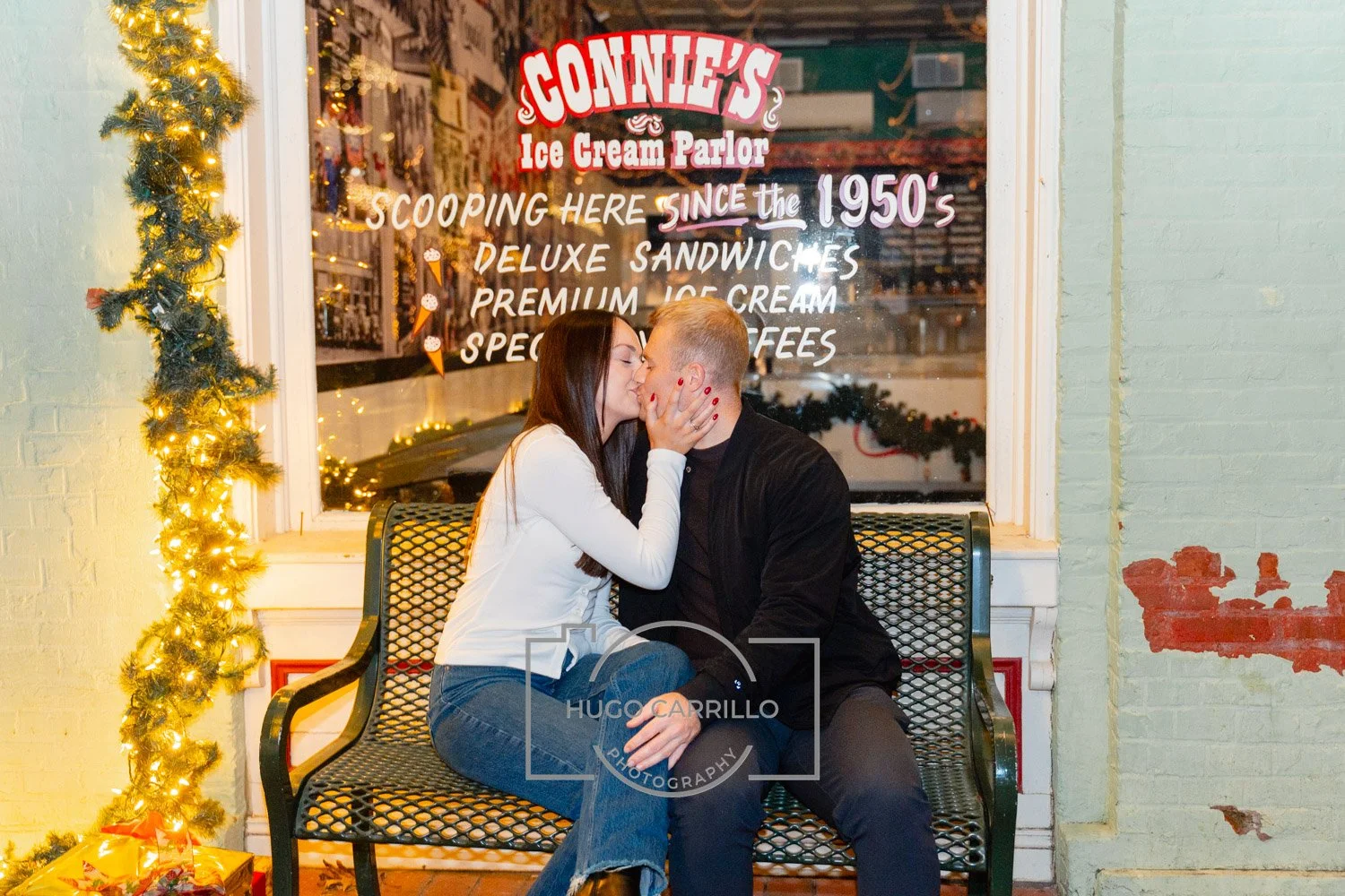 A couple sharing a kiss sitting on a bench outside Connie's Ice Cream Parlor decorated with Christmas lights and a small Christmas tree. The woman has long dark hair, white sweater, and blue jeans, while the man has short blond hair, black jacket, an