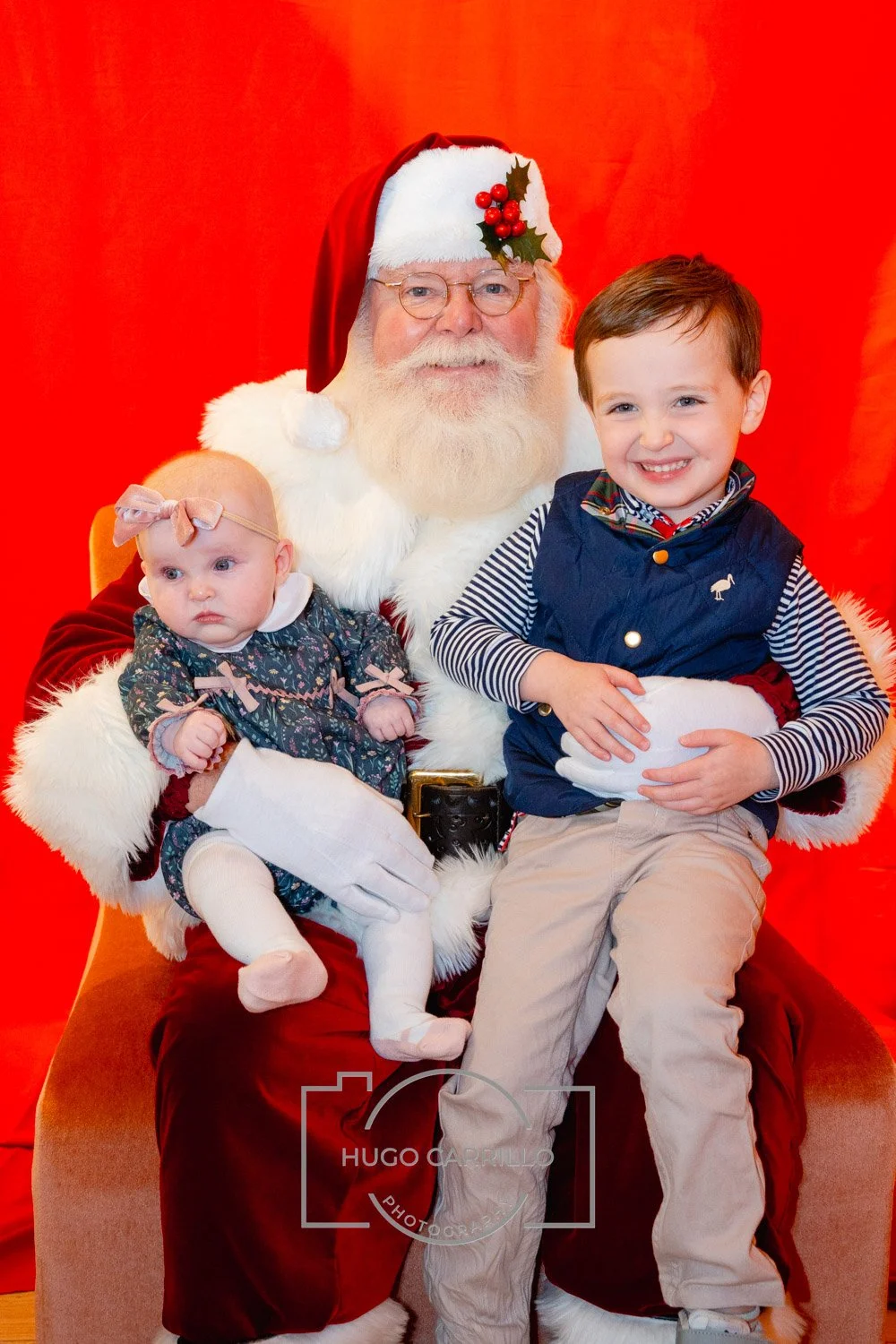 A jolly man dressed as Santa Claus sitting on a brown chair with a red backdrop, holding a baby girl dressed in a floral dress and tights, and a young boy in a navy vest and striped shirt sitting on his lap, all smiling.