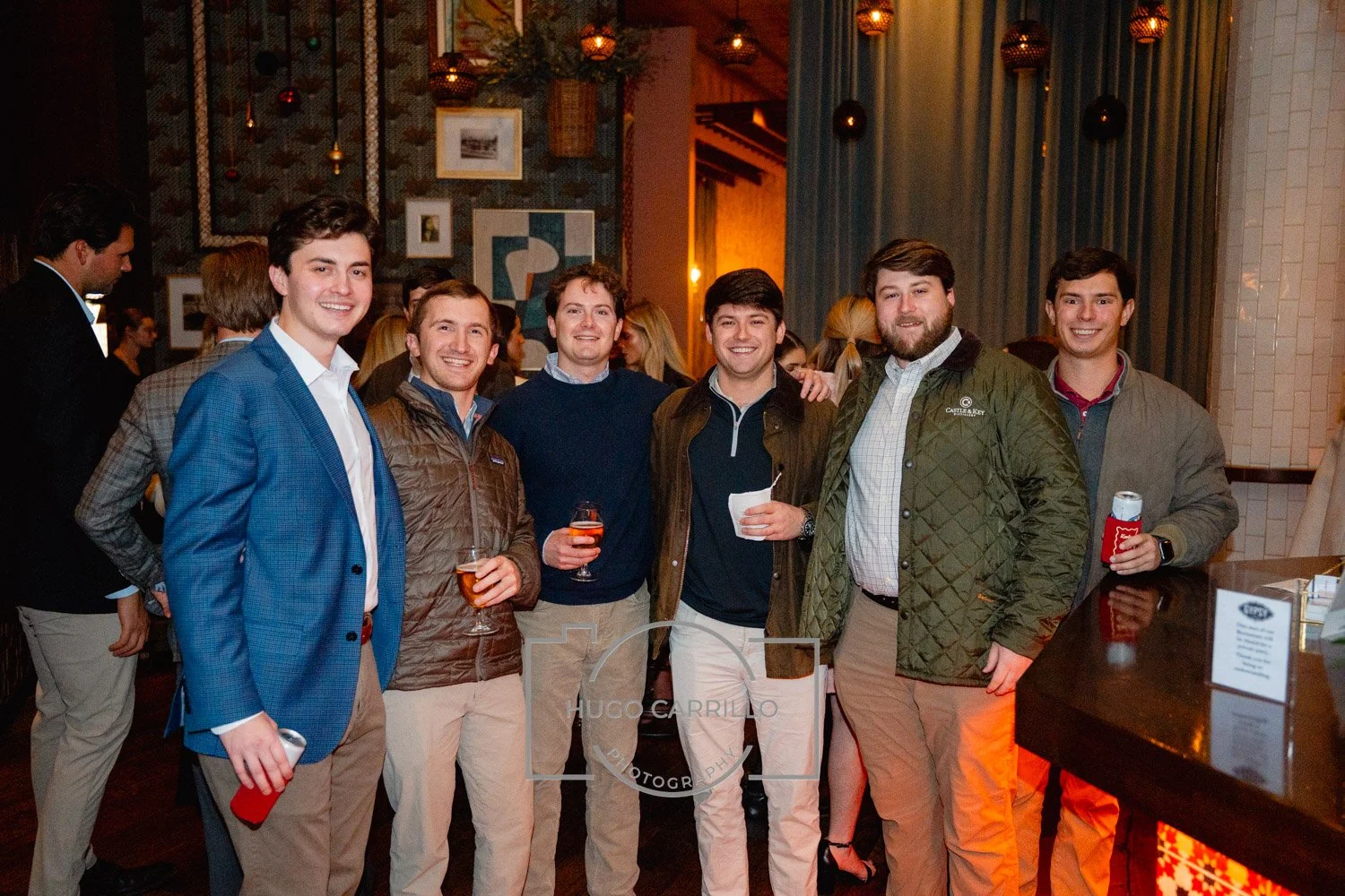 Group of six young men posing at a social event, smiling, holding drinks, in a warmly lit venue with decorative wall art and curtains in the background.