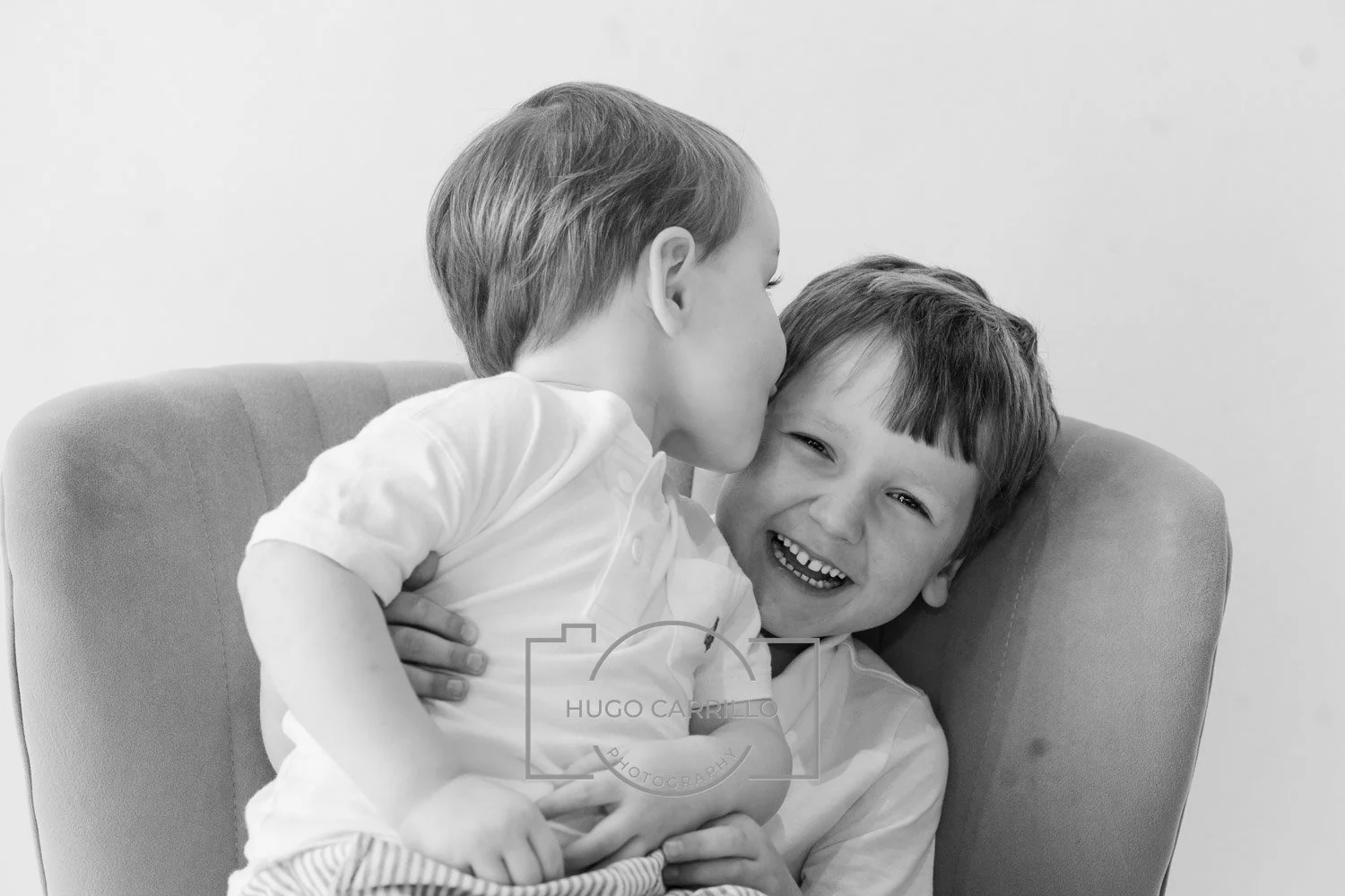 Two young boys sharing a joyful moment, one giving a kiss on the cheek to the other while sitting on an armchair.