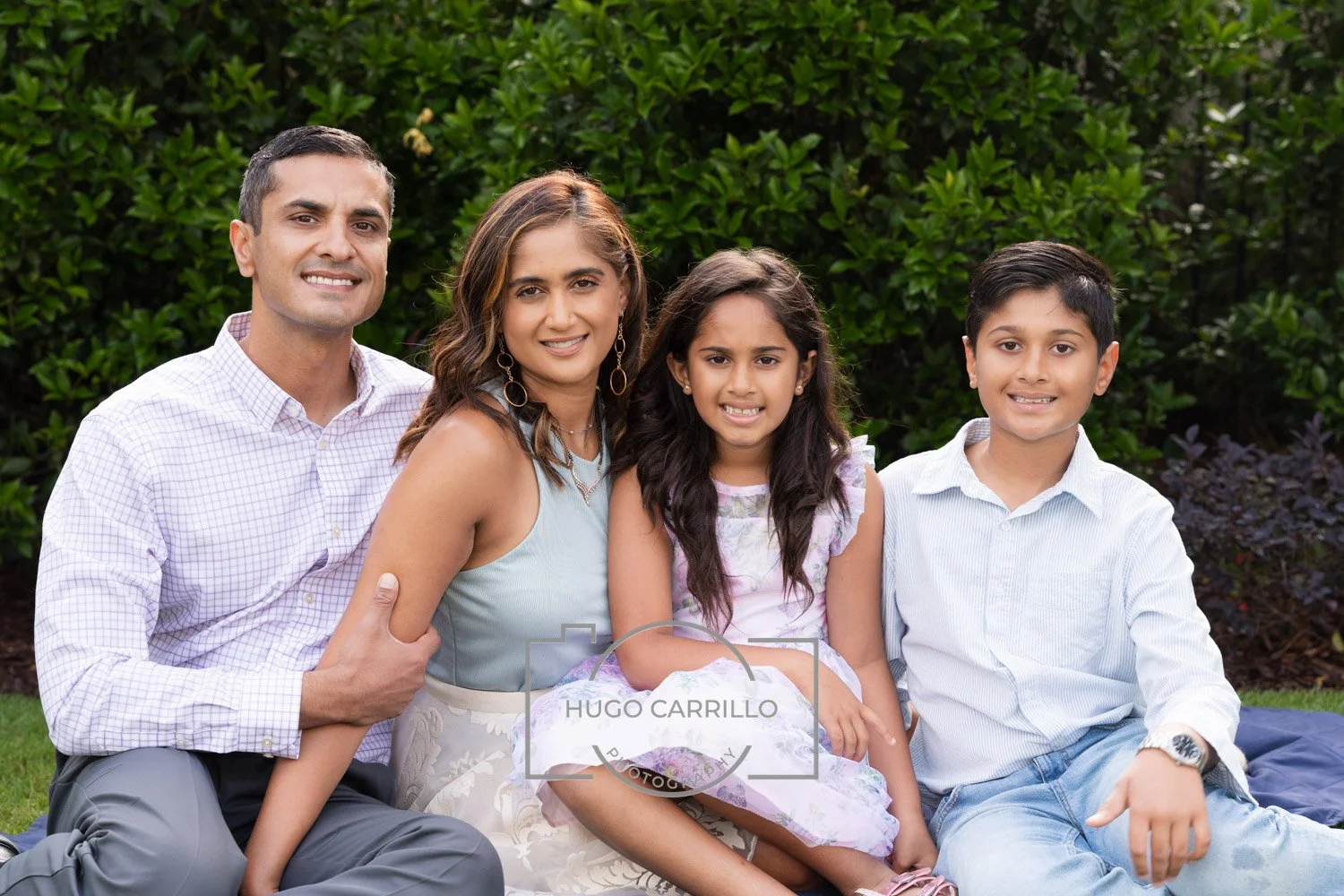 A family of four sitting outdoors on grass, with green bushes in the background. The father, mother, daughter, and son are smiling at the camera.