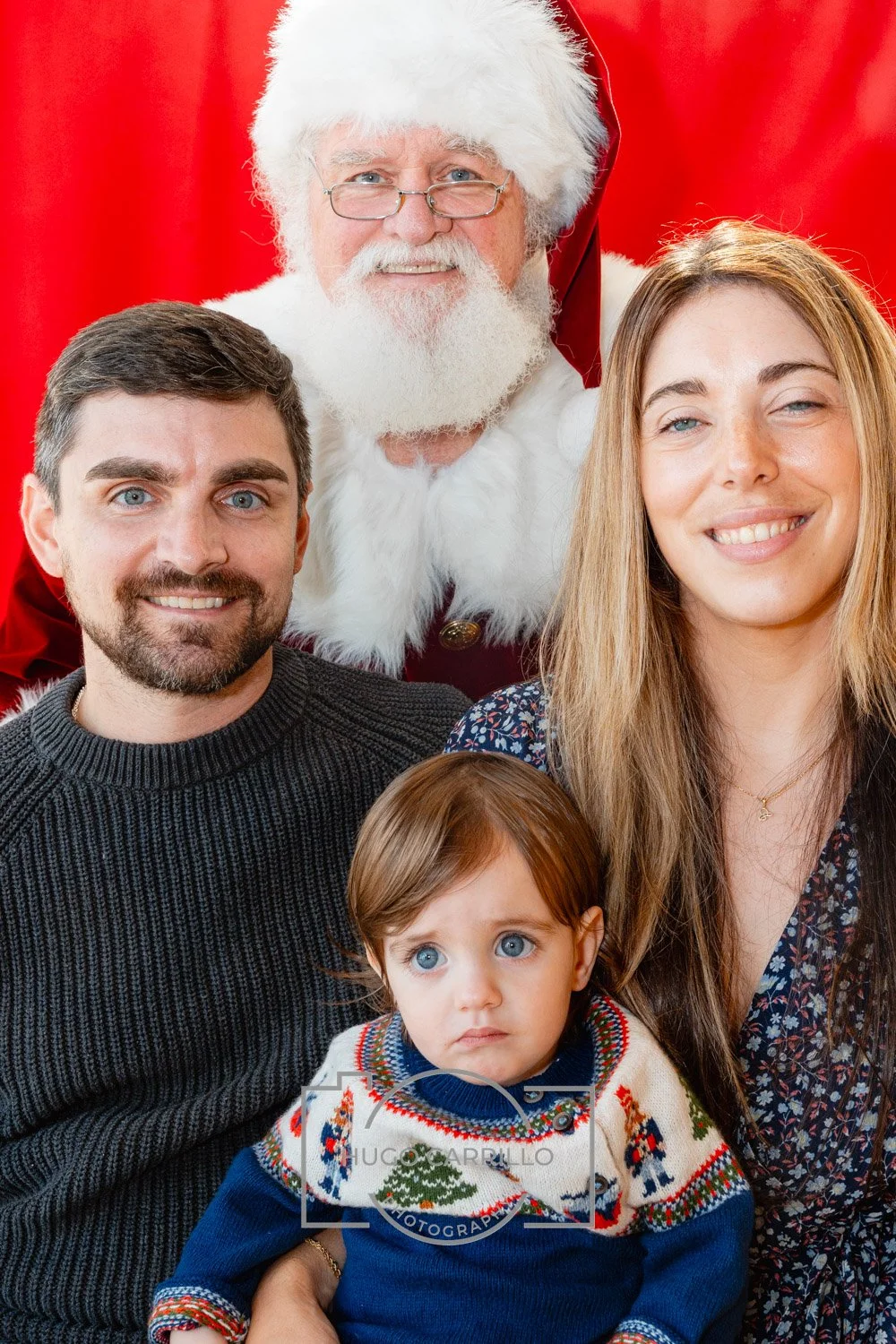 Family posing with Santa Claus for a Christmas photo, with a red background, including a young child with blue eyes in Christmas sweater.