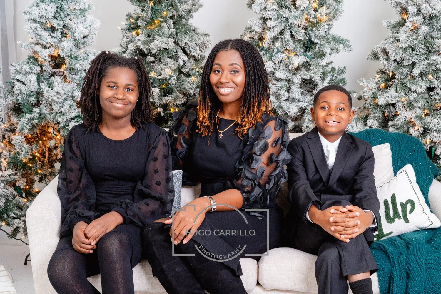 A woman with two children sitting on a white sofa in front of decorated Christmas trees, smiling for a holiday photo.
