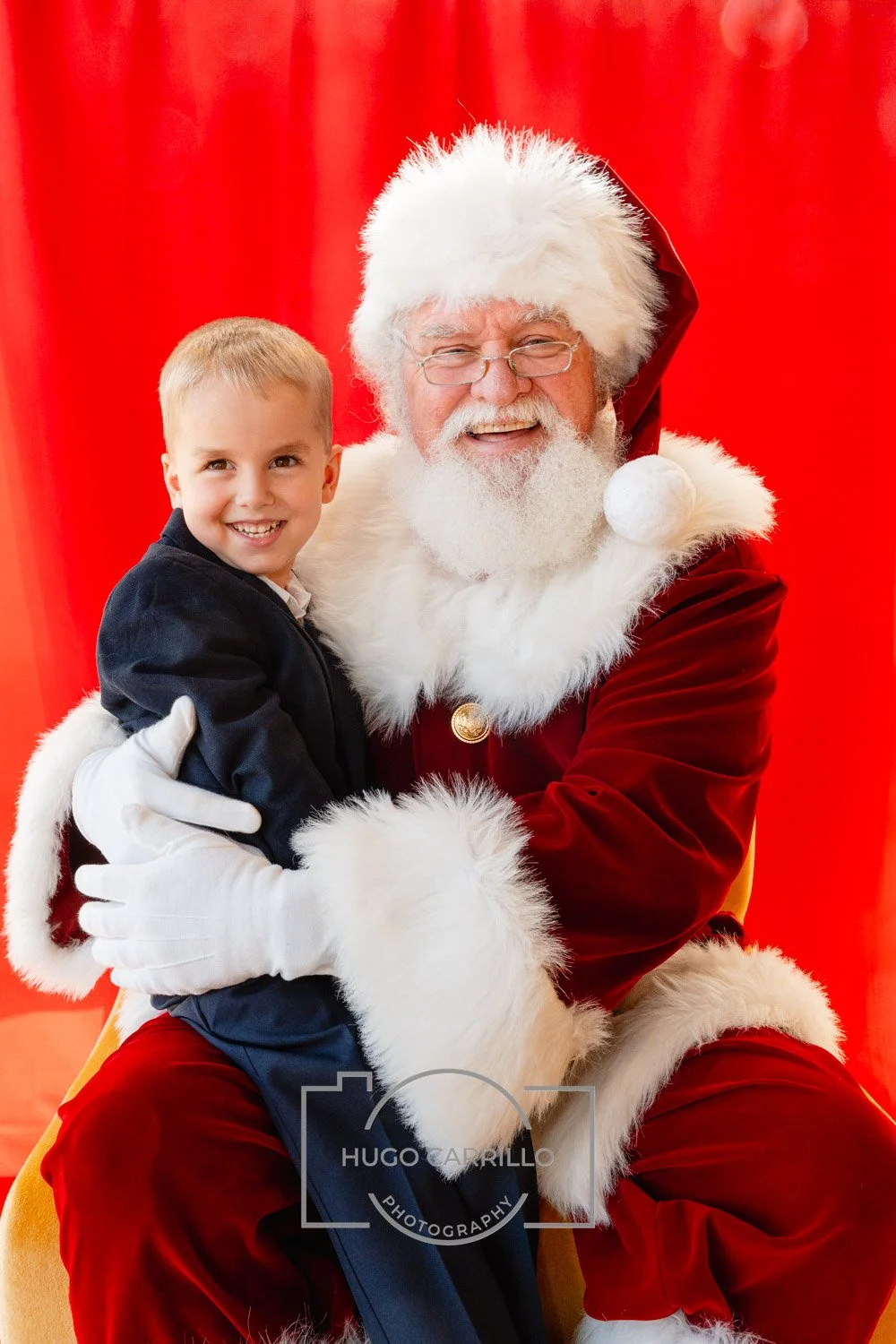 A young boy in a black suit sitting on Santa Claus's lap, both smiling happily. Santa is dressed in a traditional red velvet suit with white fur trim, glasses, and a red hat with a white pompom. The background is red.