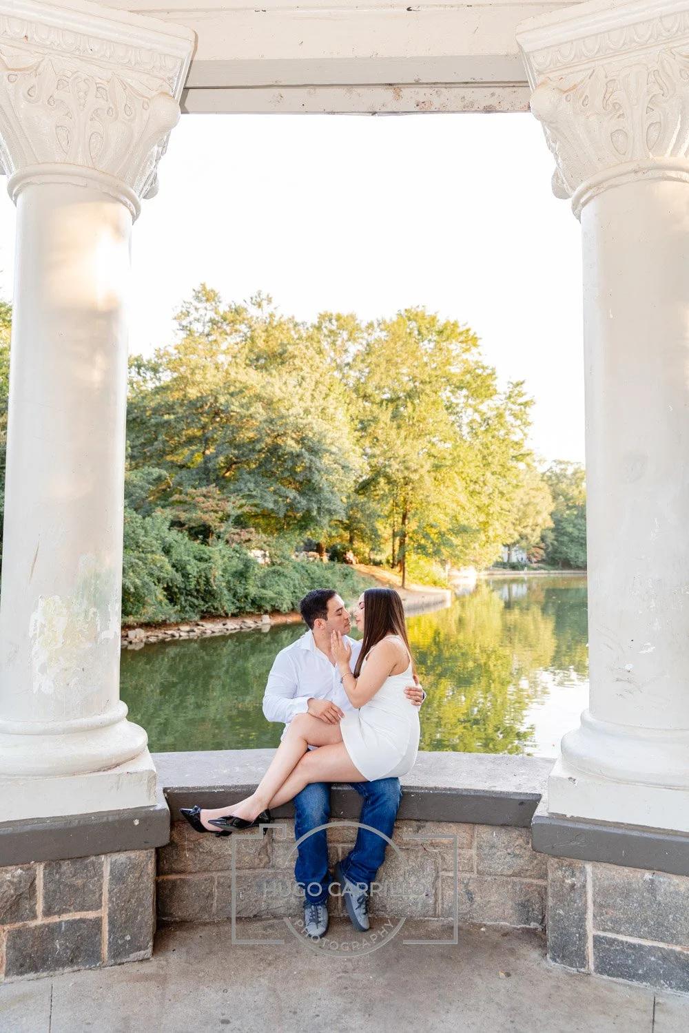 A couple sitting on a stone ledge under a white pavilion with columns, near a body of water with trees in the background, during sunset.