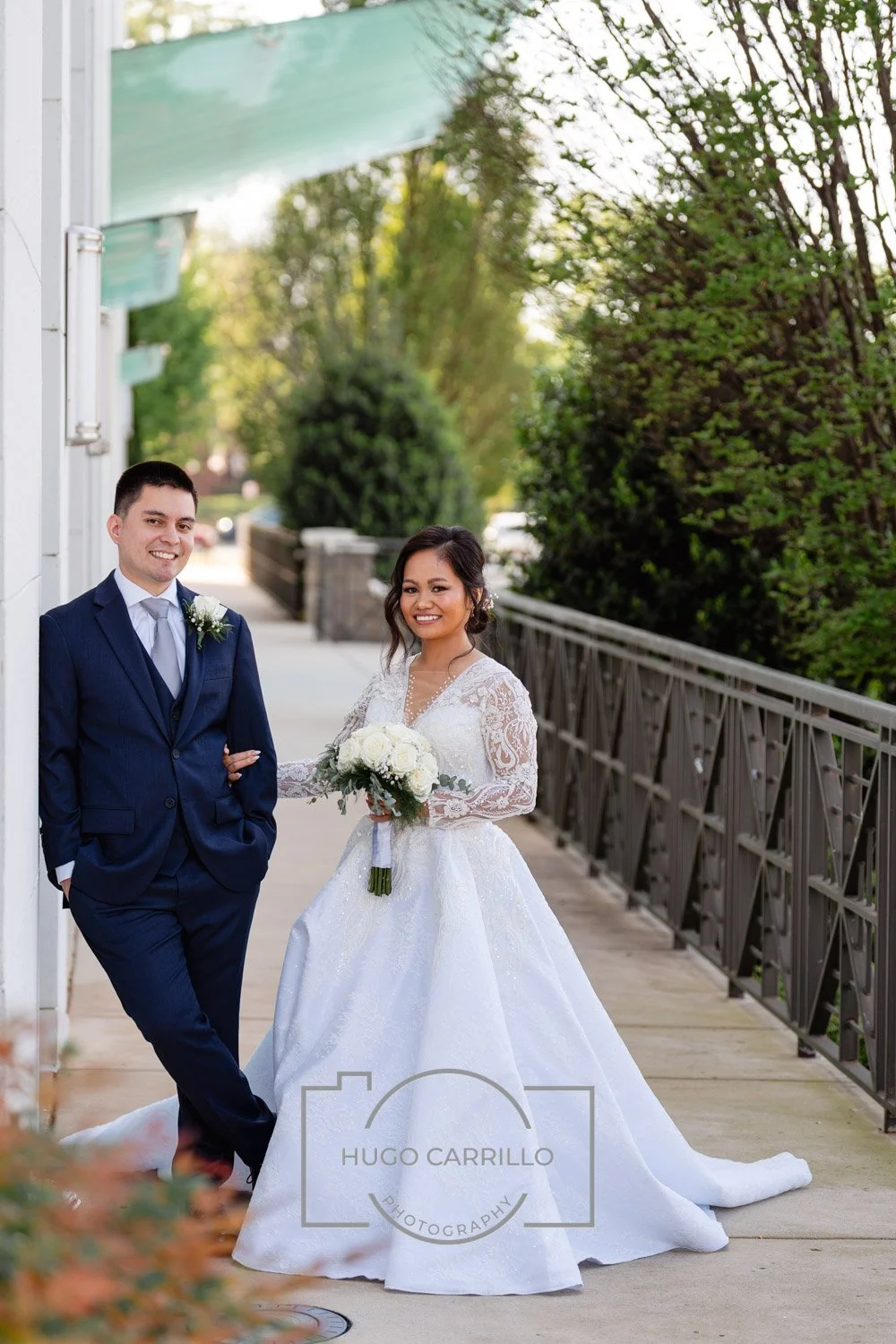 Happy bride in a white wedding gown holding a bouquet and groom in a navy suit with a boutonnière, standing outdoors on a sidewalk with greenery and a bridge railing in the background.