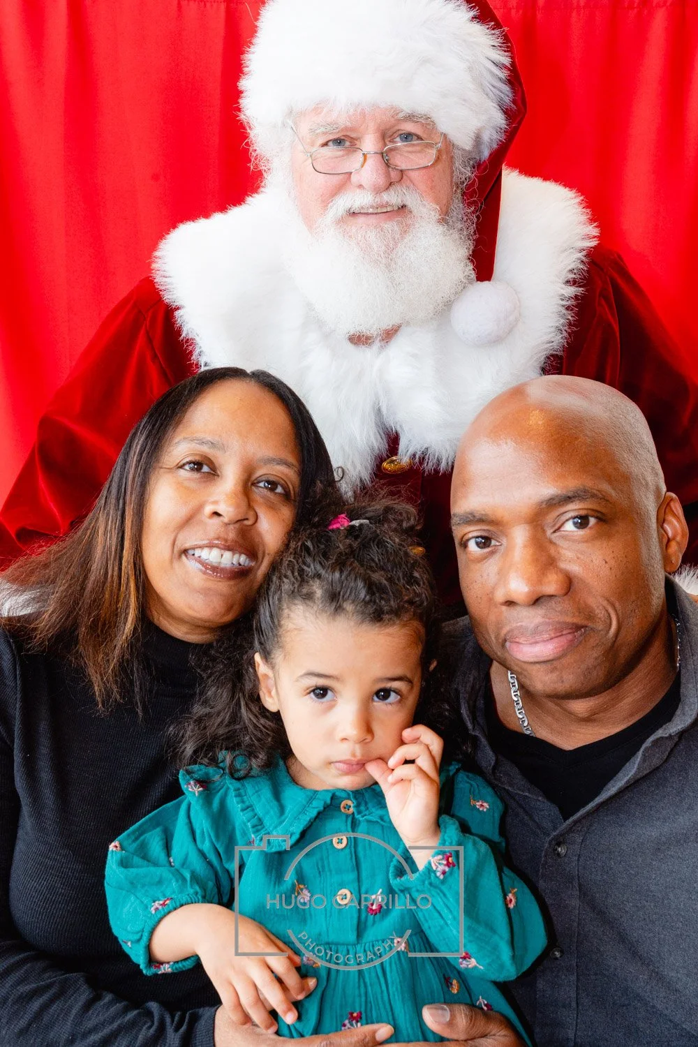 A family of three with a young girl, posing with Santa Claus in front of red curtains. Santa is dressed in a traditional red suit with white trim, glasses, and a white beard. The family members are smiling, except for the girl who looks thoughtful.