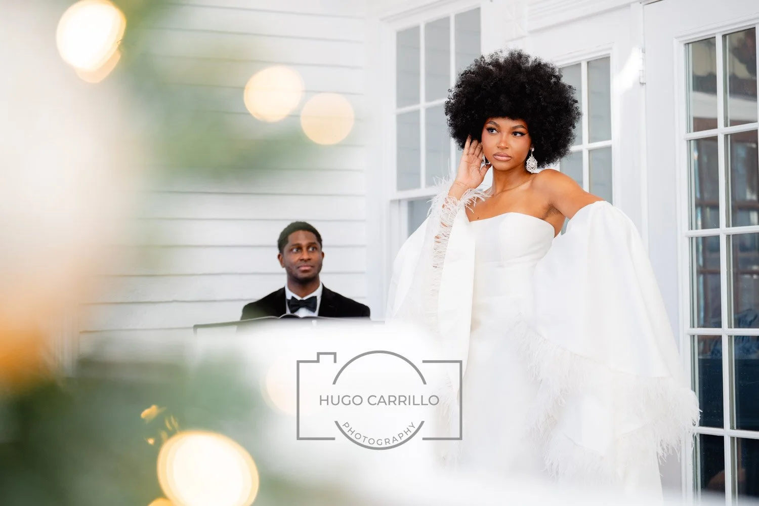 A woman in a white wedding dress with puff sleeves, wearing large earrings, stands indoors near a large window, touching her ear, while a man in a tuxedo sits behind her, looking at her, during a wedding ceremony or celebration.