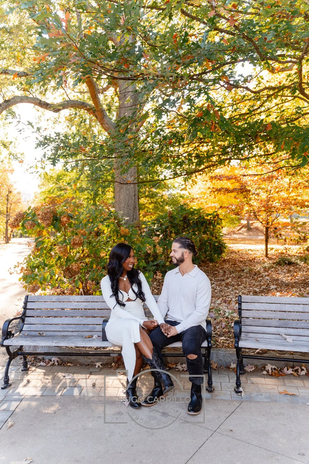A man and woman sitting on a park bench, holding hands, smiling and looking at each other on a sunny autumn day with vibrant fall foliage in the background.