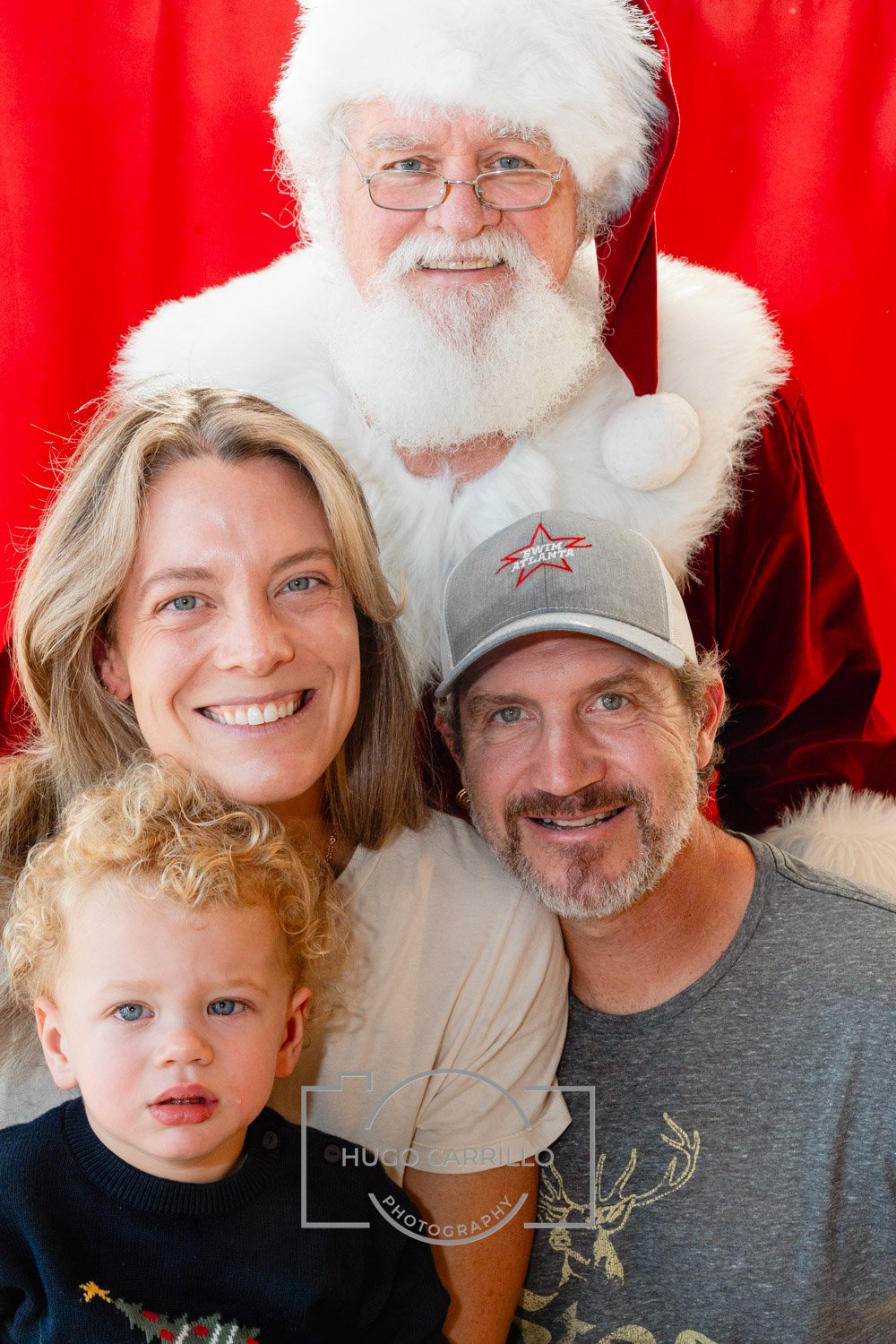 A family of three posing with Santa Claus in front of a red backdrop. The woman has shoulder-length blonde hair, the man wears a gray cap, and the young boy has curly blonde hair and is wearing a Christmas sweater.