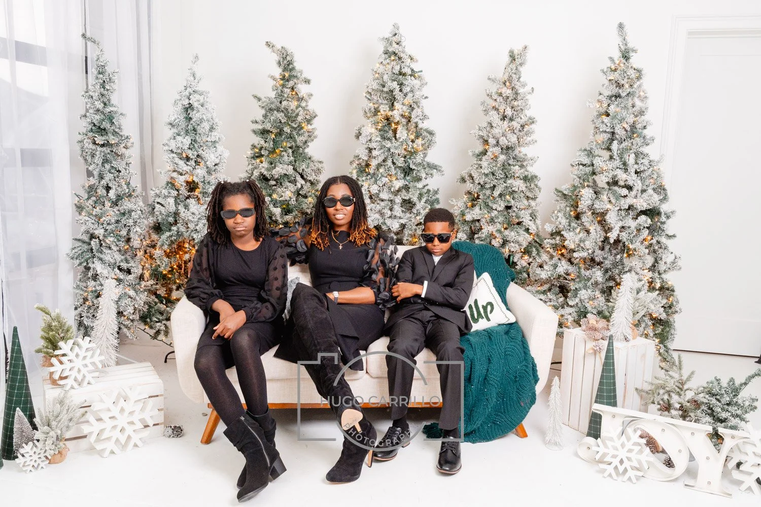 A woman and two children sitting on a white sofa with a Christmas tree backdrop. All are wearing black clothes and sunglasses, with Christmas decorations and snowy trees around them.