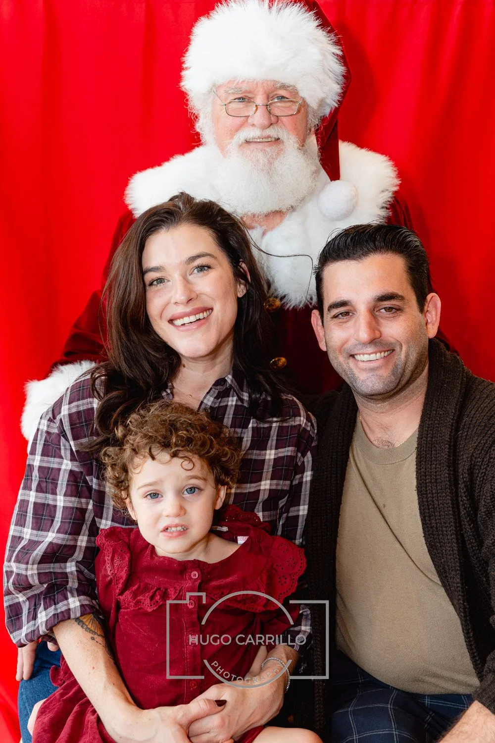 A family of four posing with Santa Claus against a red backdrop. The family includes a woman with long brown hair, a man with short dark hair, a young girl with curly brown hair, and a toddler with curly red hair. Santa is dressed in his traditional 
