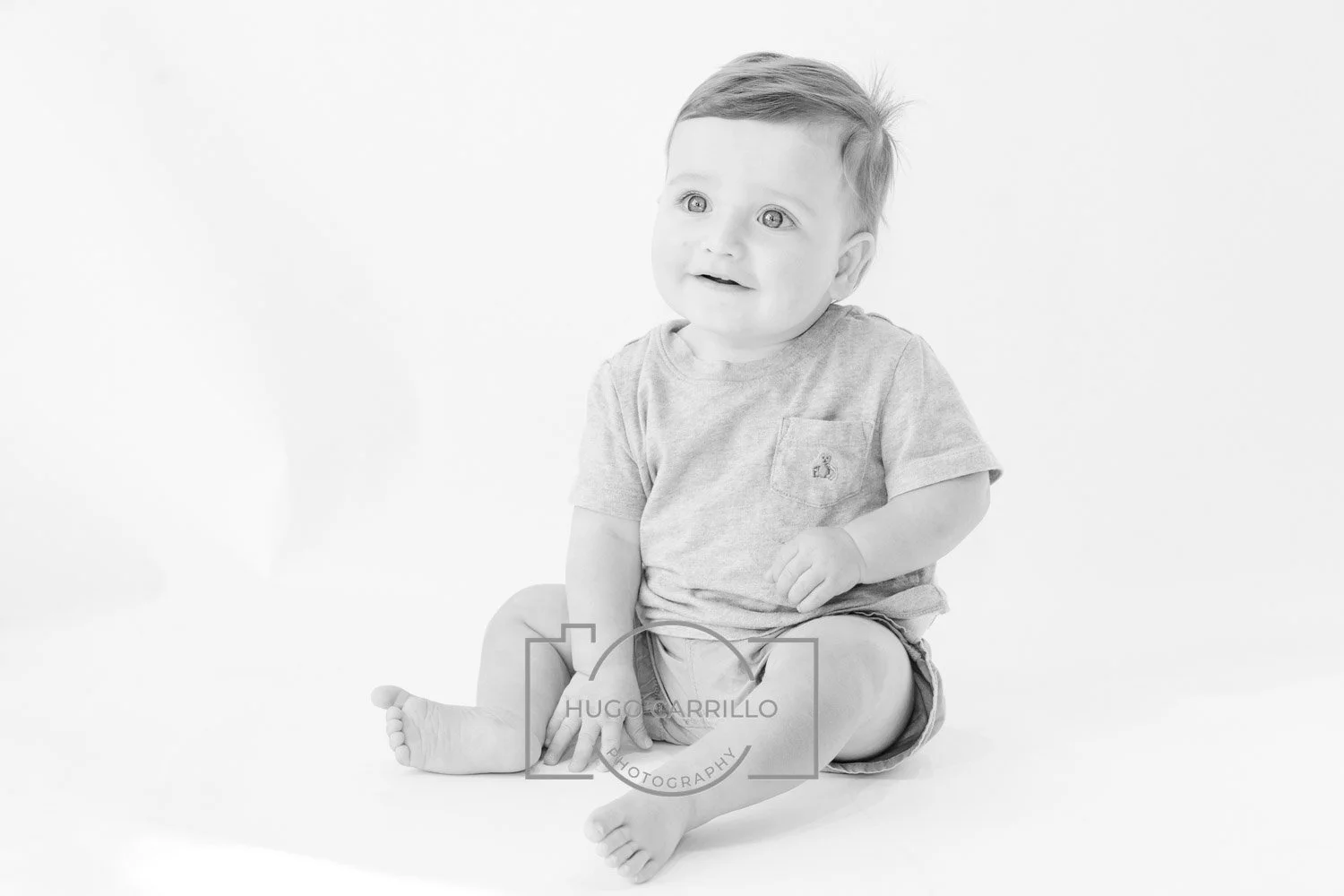 Black and white photo of a happy baby sitting on the floor against a plain background. The baby has short hair, big eyes, and is wearing a T-shirt and shorts.