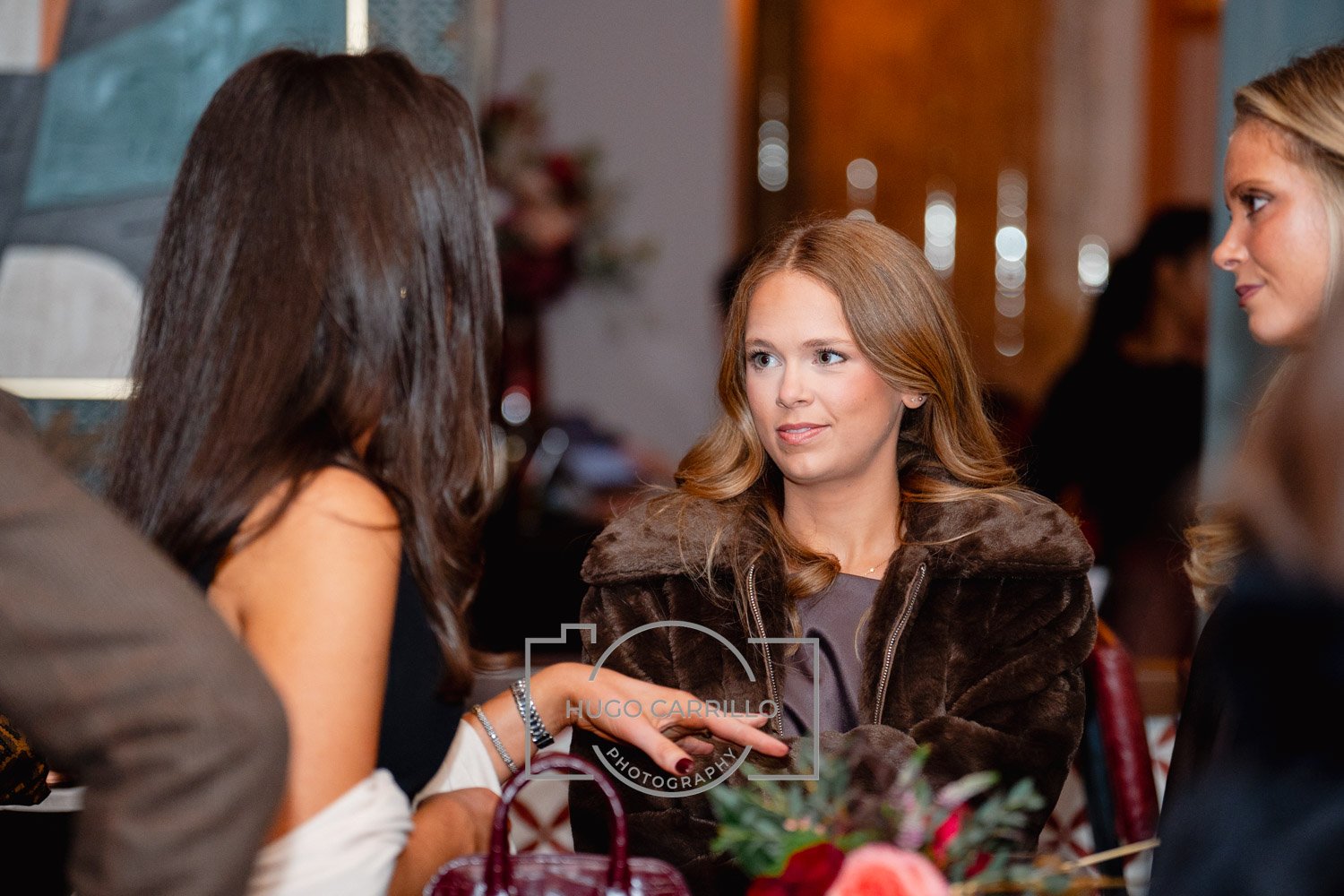 Three women engaging in conversation at a social event, with a woman in the center wearing a brown fur coat and a woman on the left gesturing with her hand, in a warmly lit room with blurred background.