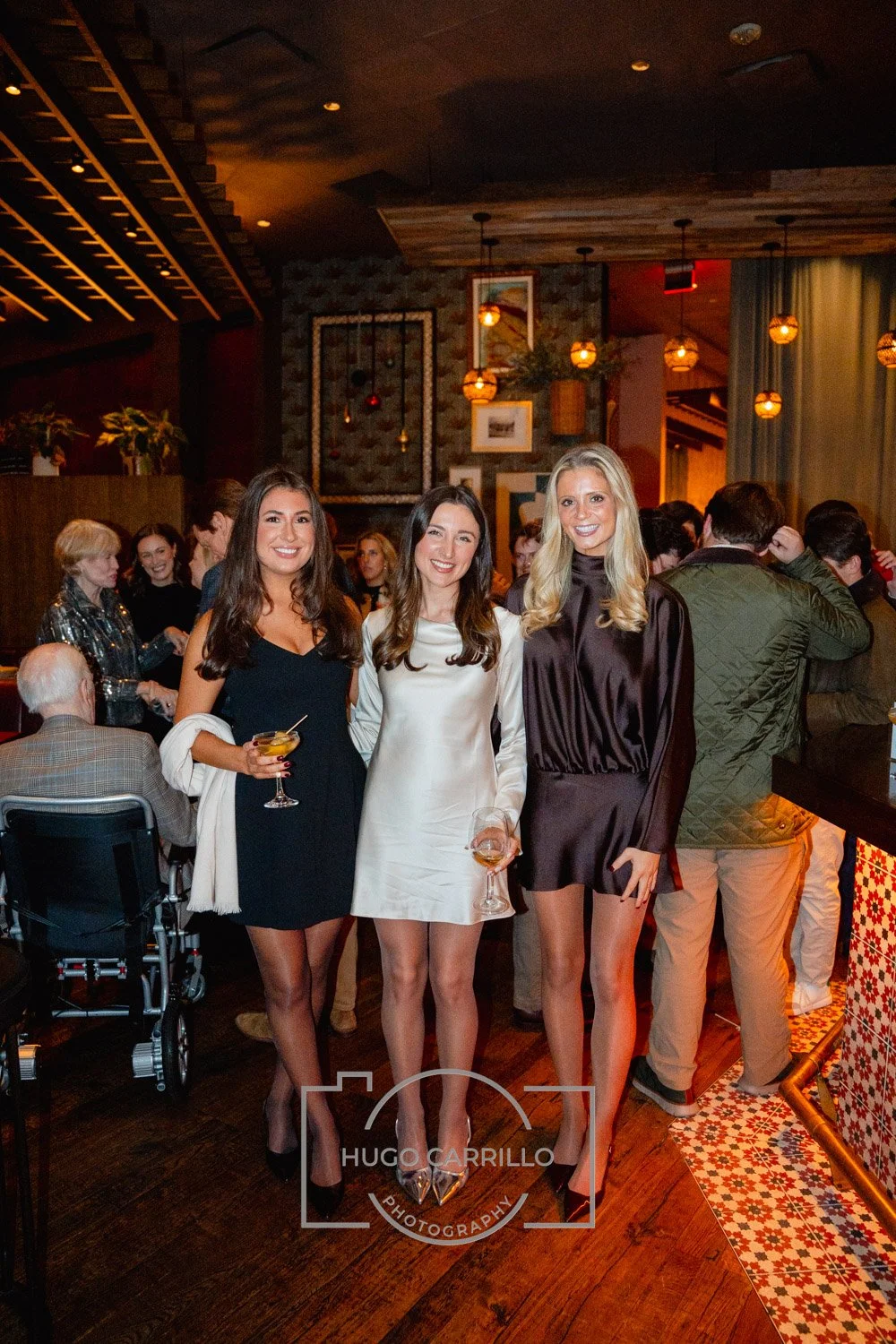 Three women in elegant dresses smiling and holding drinks at a social gathering in a warmly lit restaurant or bar.