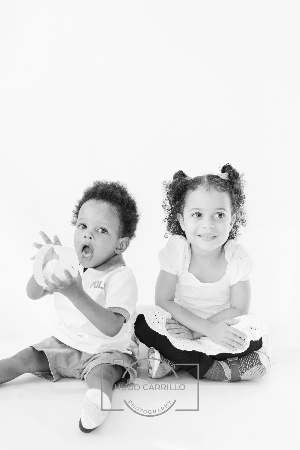 Black and white photo of two young children sitting on the floor with plain white background. The boy on the left has curly hair and is holding a round object, possibly a toy, while the girl on the right has pigtails and is smiling with her hands res