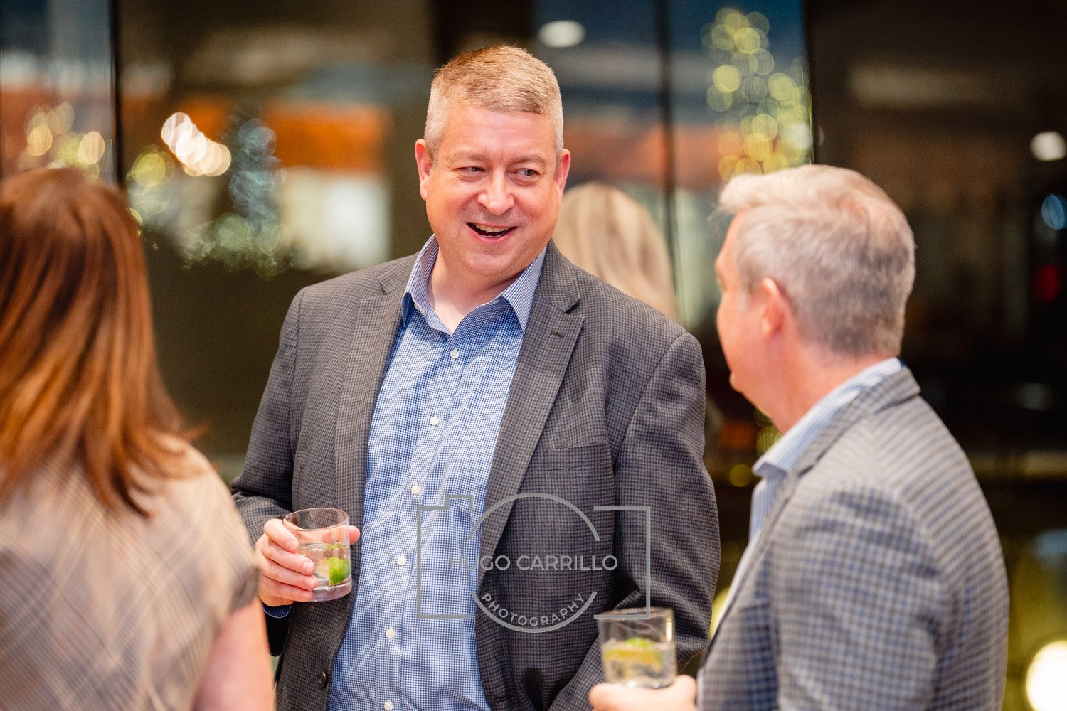 A group of three people having a conversation at an event. The man in the center, with short gray hair, is smiling and holding a drink with lime. The man on the right, with gray hair, is also holding a drink with lime. The woman on the left has reddi