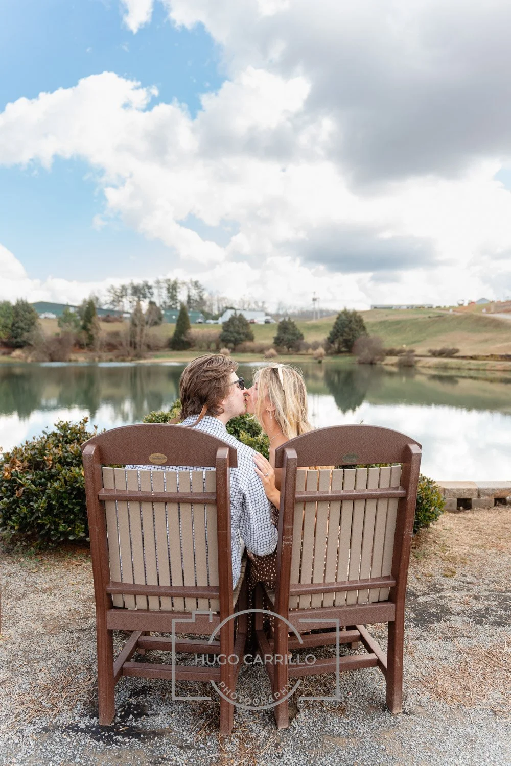 A couple sharing a kiss while sitting on outdoor chairs near a lake with trees and cloudy sky in the background.