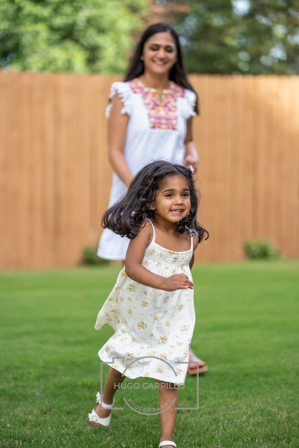 A girl with dark curly hair running on a grassy yard, wearing a white dress with floral print, while a woman in a white embroidered dress watching her in the background.