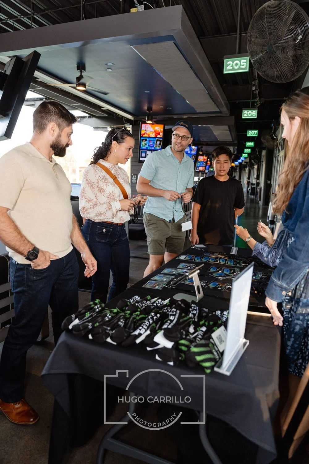 Group of five people standing around a table with merchandise, including socks and digital devices, inside a sports stadium or arena, with digital displays and a large fan overhead.