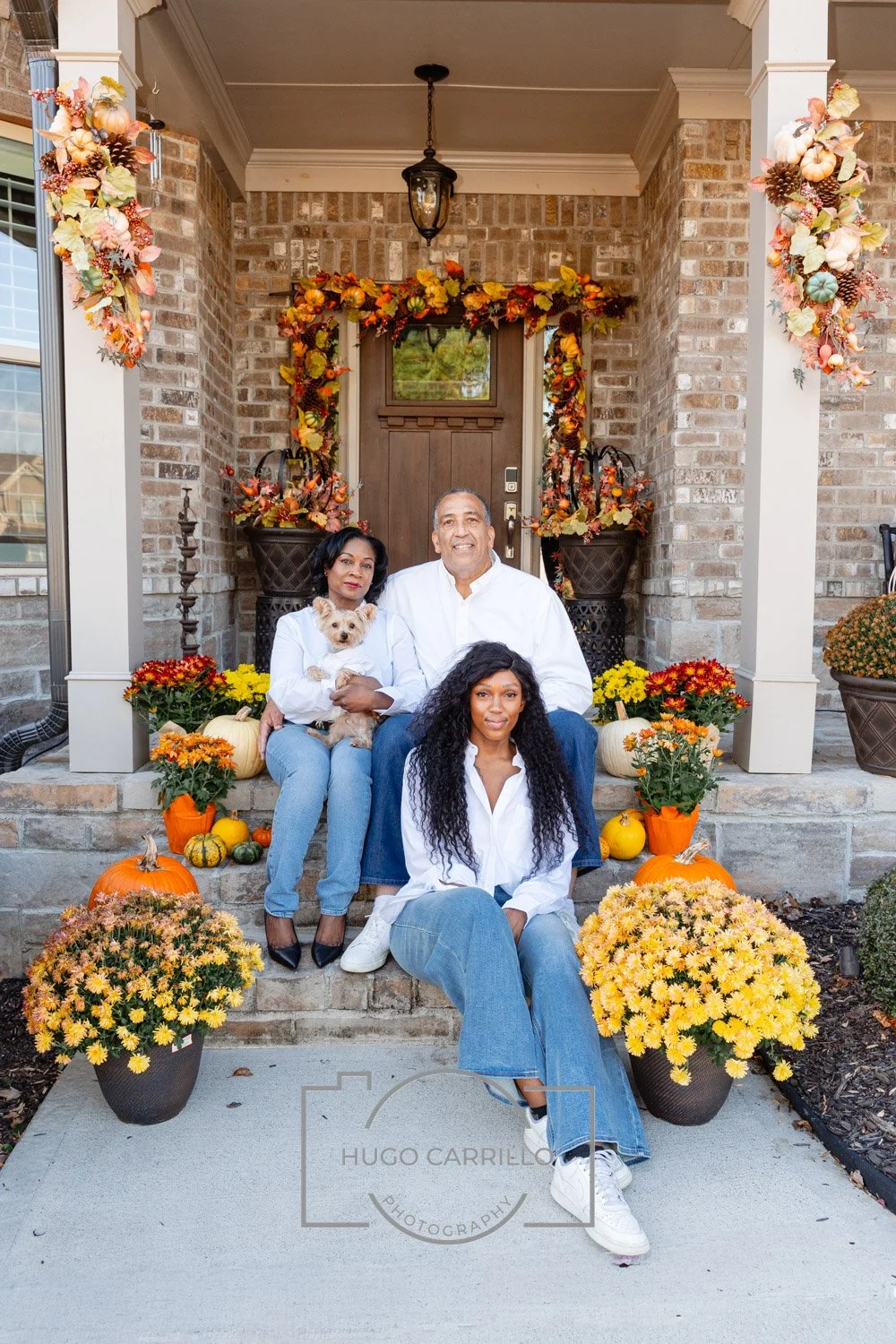 A family of three women, one man, and a dog sitting on the steps of a house decorated with fall and Halloween themed pumpkins, flowers, and autumn leaves around the porch.