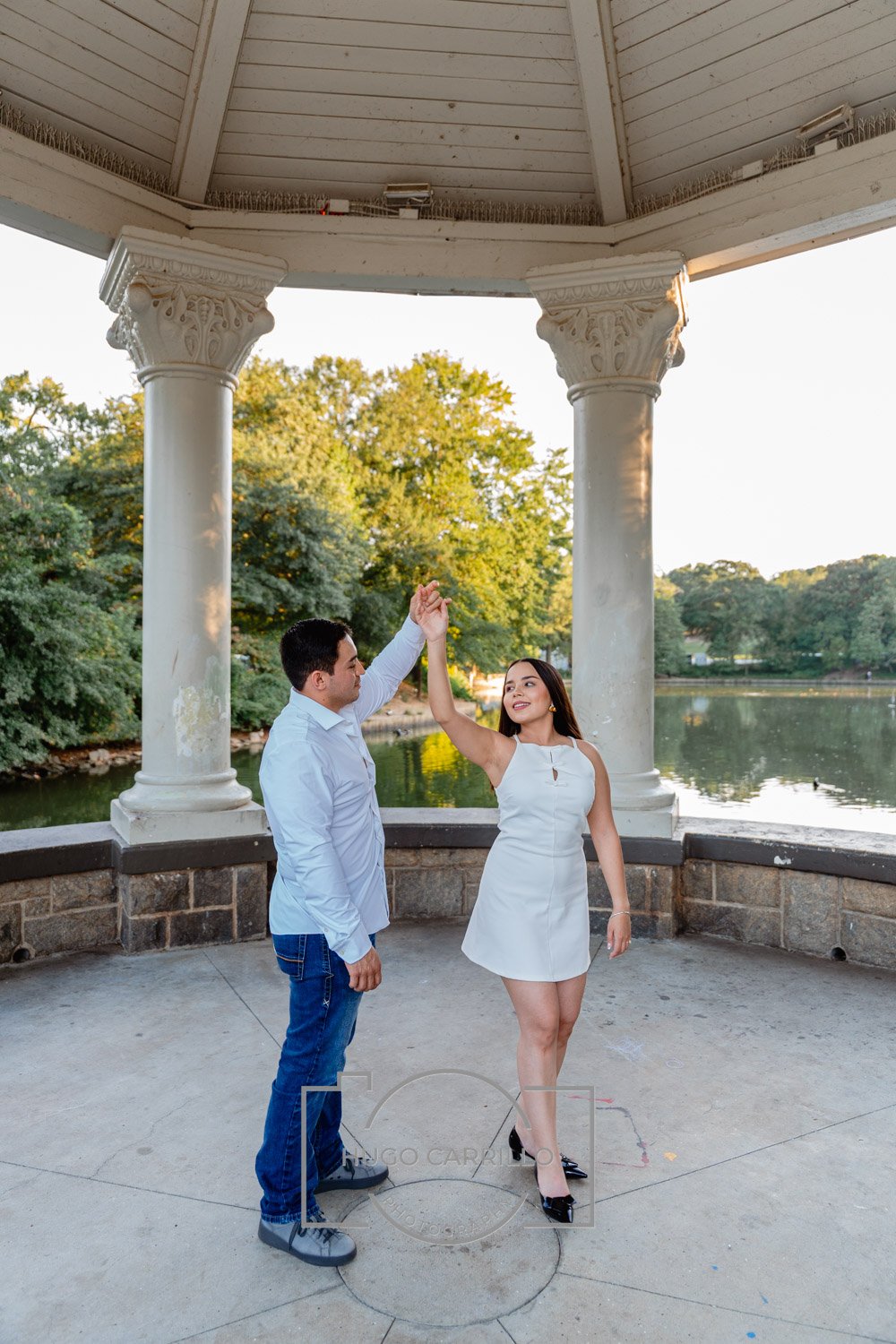 A couple dancing together in a gazebo near a lake, with trees and water in the background during sunset.
