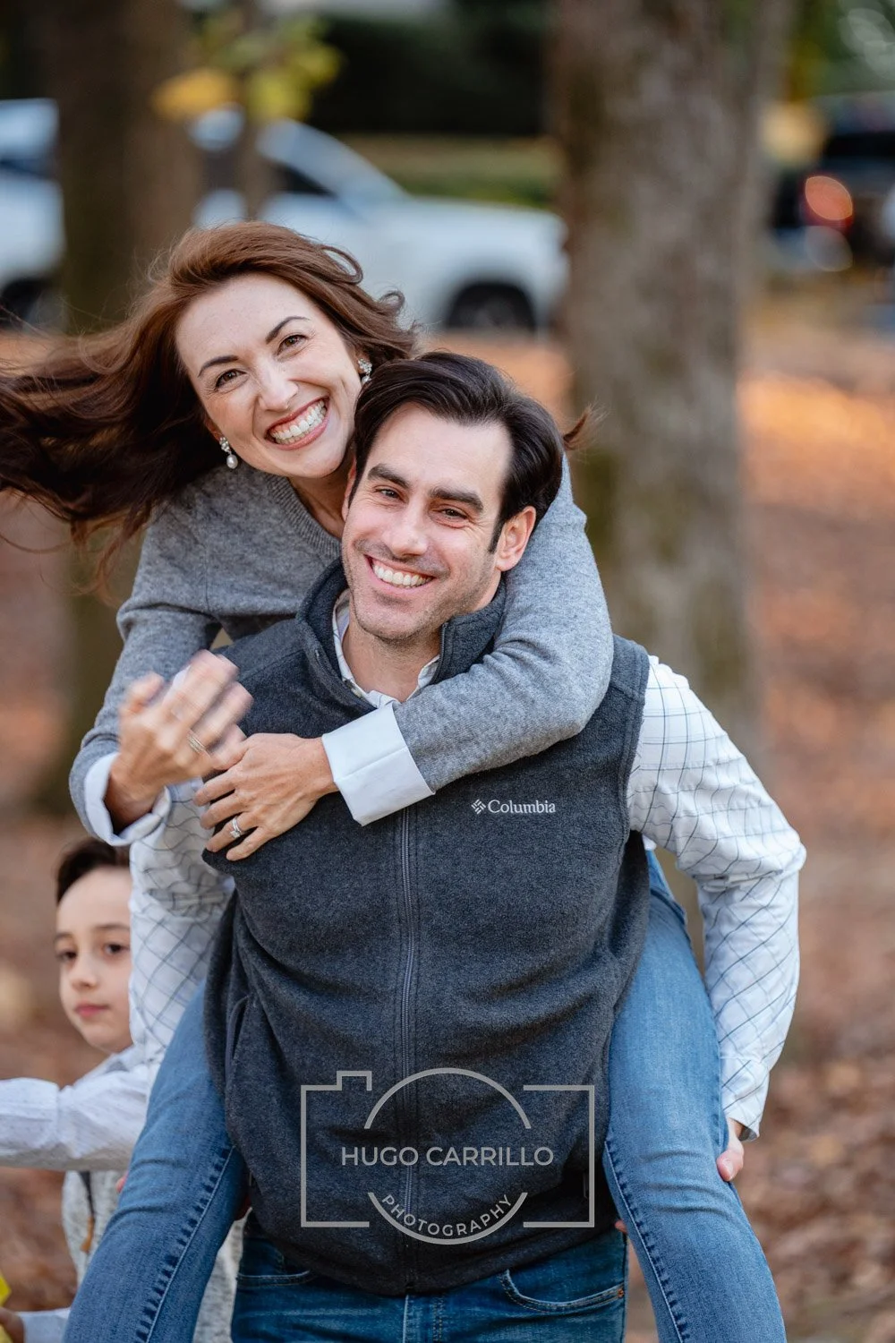 A smiling woman with long brown hair and earrings piggybacking on a man wearing a Columbia vest, outdoors in autumn, with a child partially visible in the background.