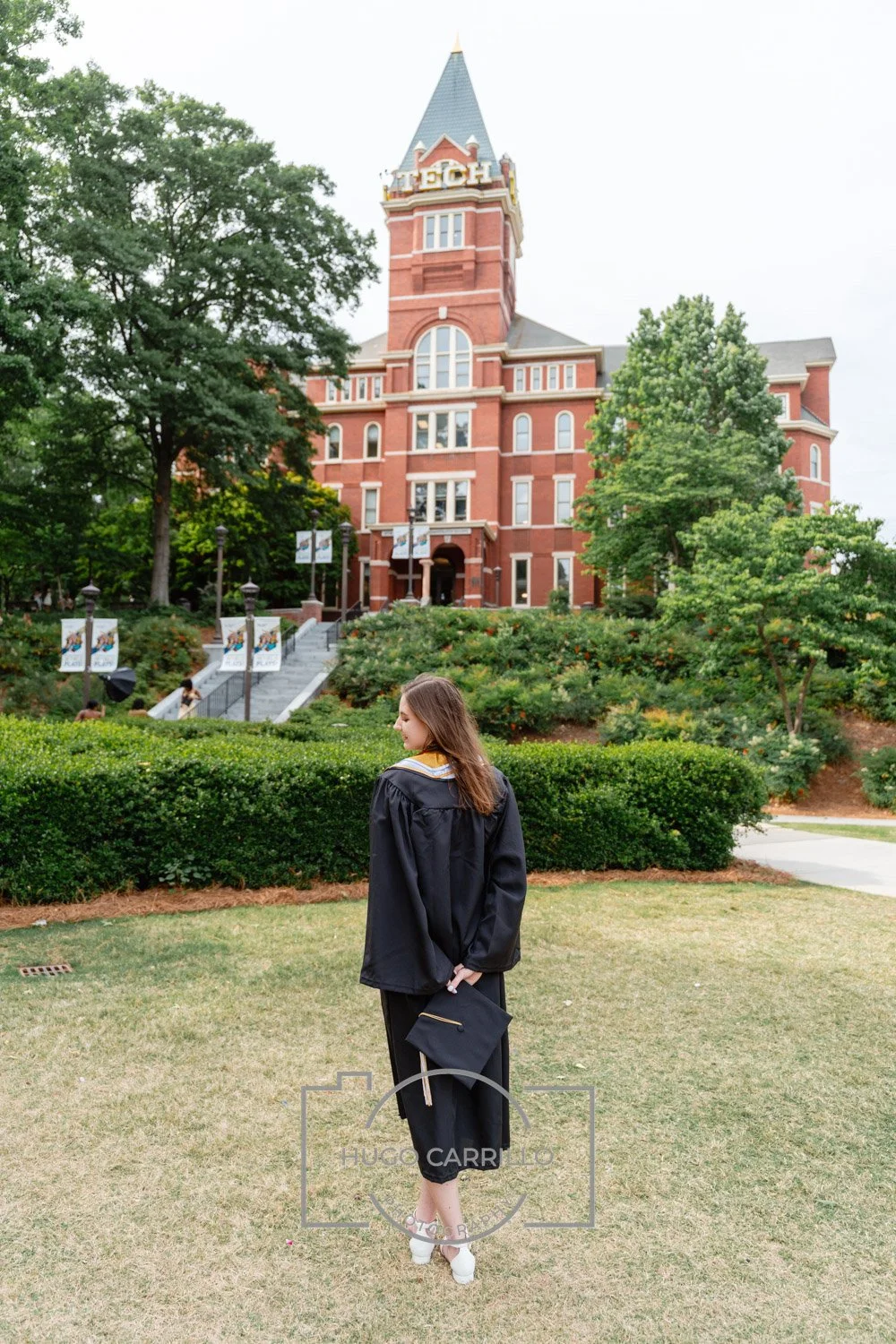 A young woman in a graduation gown holding a cap stands on a grassy area in front of a brick building with a tower that has the word 'TECH' on it. The scene is surrounded by trees and bushes.