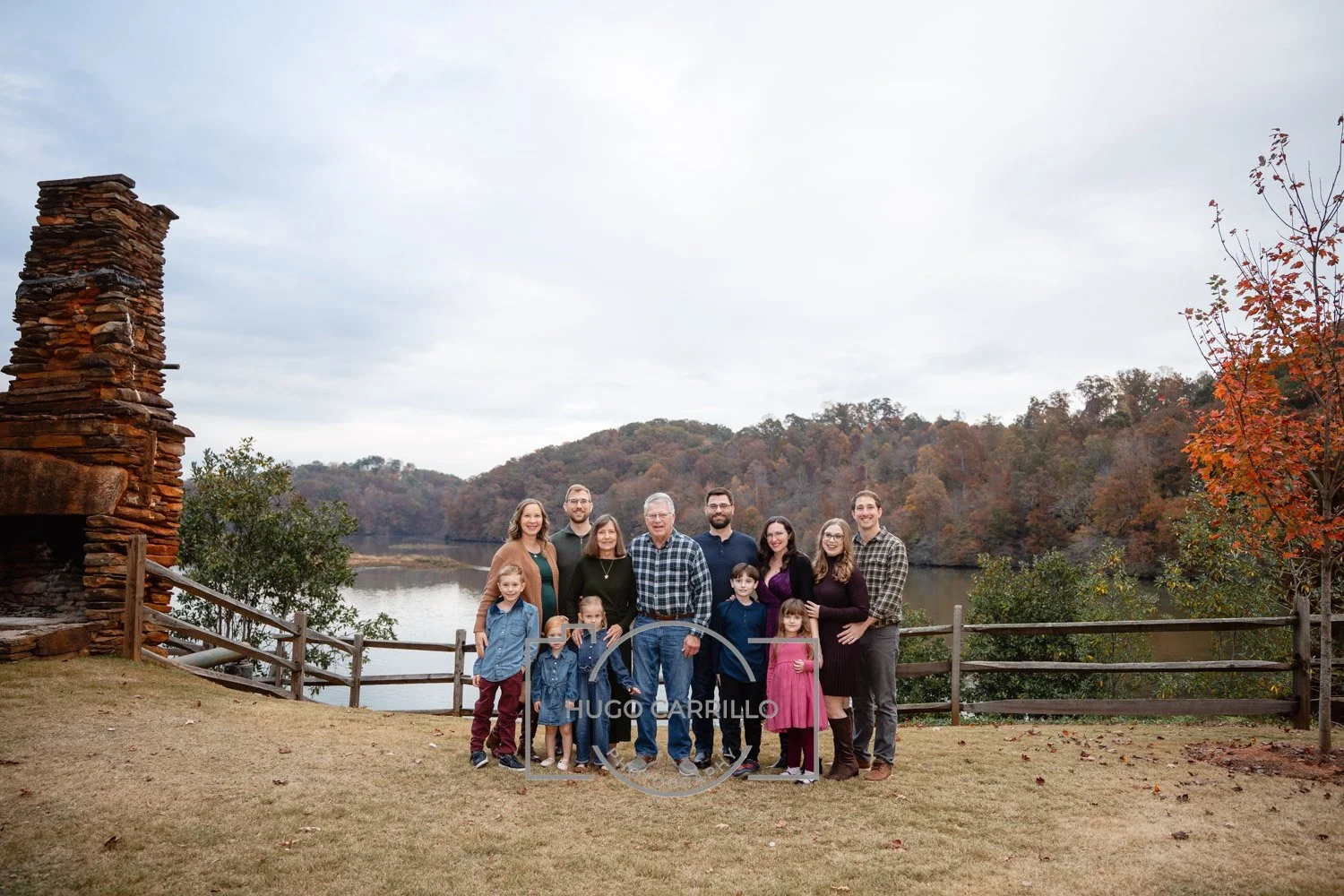A group of eleven people, including adults and children, standing on grass near a lakeside with trees and hills in the background during autumn.