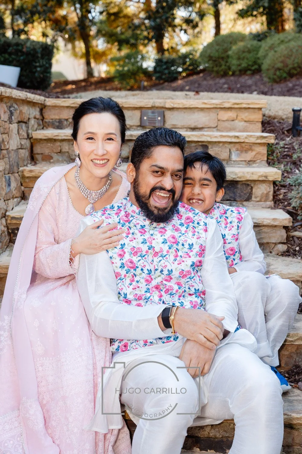 A cheerful family of three dressed in traditional Indian clothing, sitting on stone steps outdoors with trees in the background. The woman is in a pink lace dress with jewelry, the man is in a white outfit with a floral vest, and the child is in whit