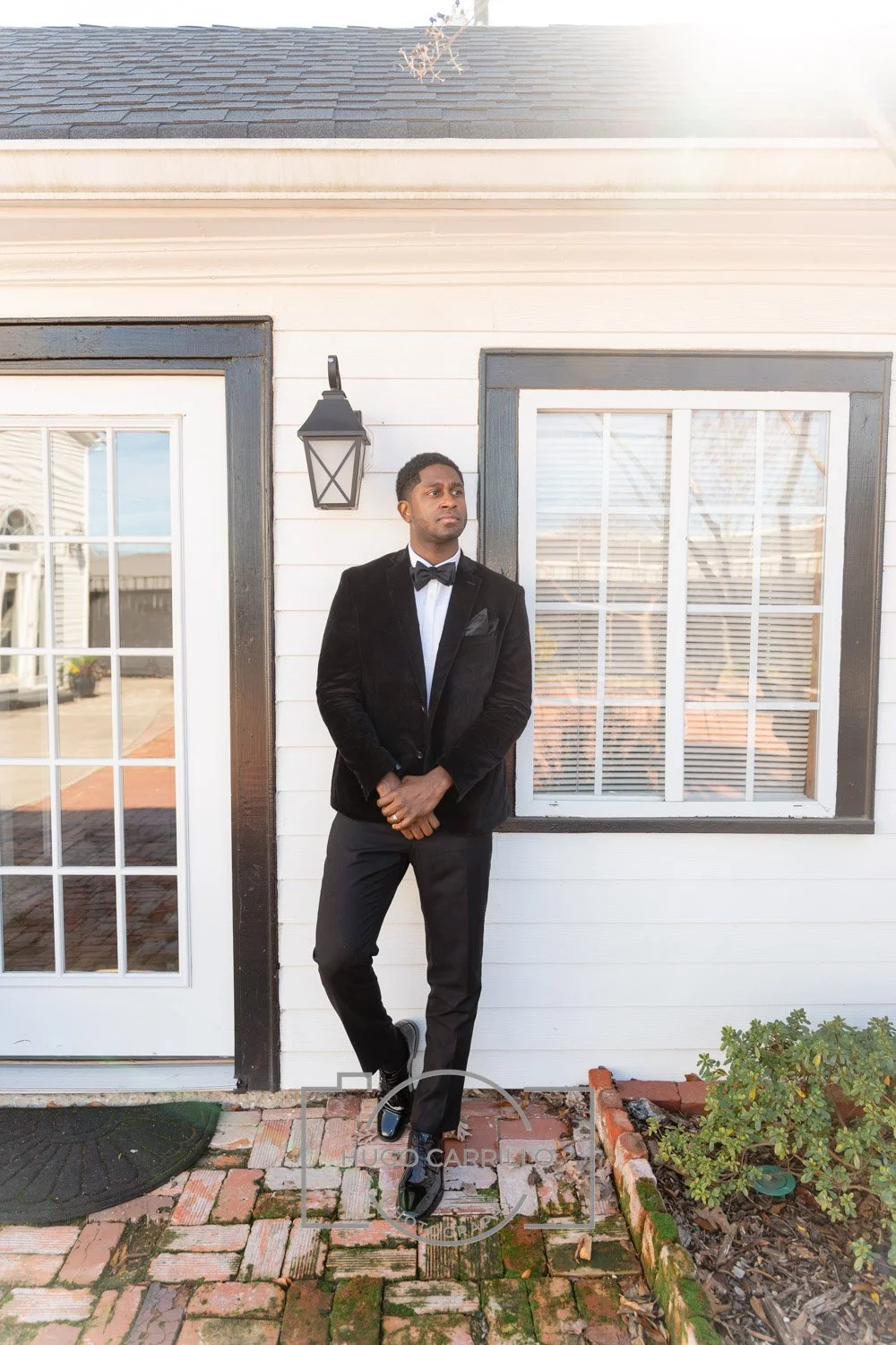 A man in a black tuxedo stands outside a white house with black trim, near a door with glass panes and a window with blinds, in a garden with brick pavers and plants.