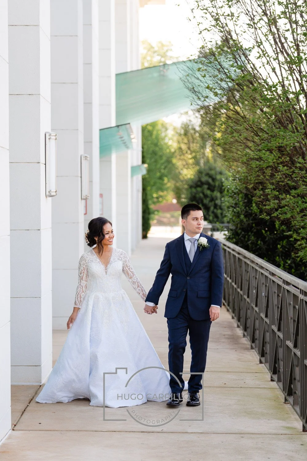 A bride and groom walking hand in hand outdoors on a paved walkway. The bride is dressed in a white lace wedding gown, and the groom is in a dark blue suit with a light blue shirt and tie, both smiling and holding hands.