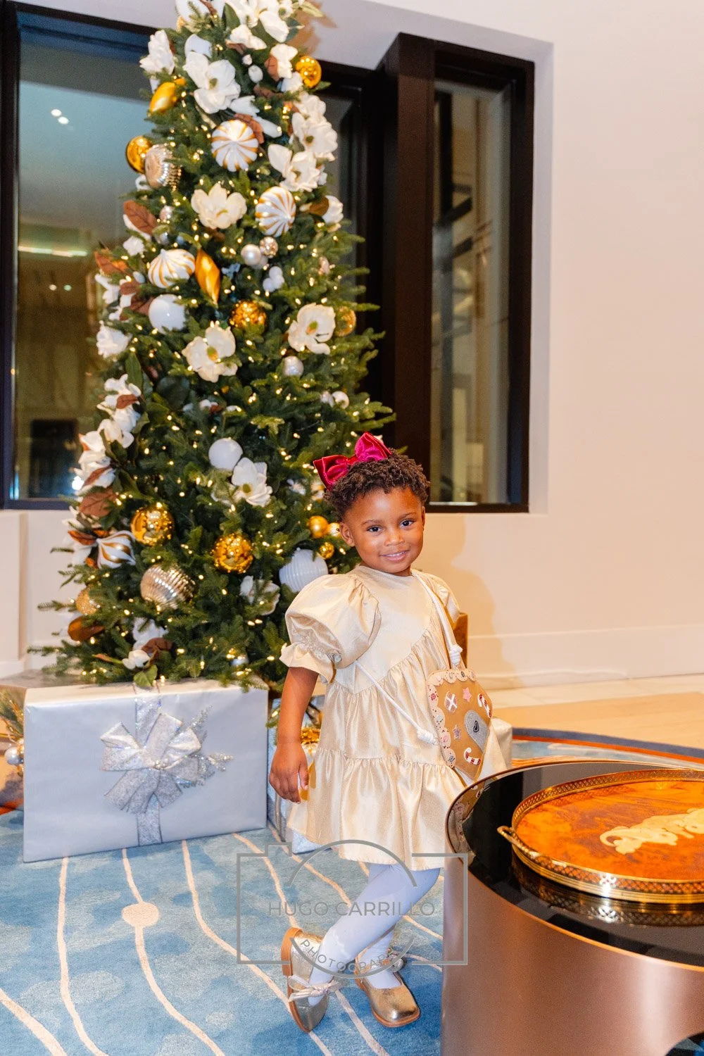 A young girl in a gold dress with puffy sleeves and a large red bow headband, standing in front of a decorated Christmas tree with white, gold, and brown ornaments, next to a large Christmas gift wrapped in silver paper. She is smiling and holding a 