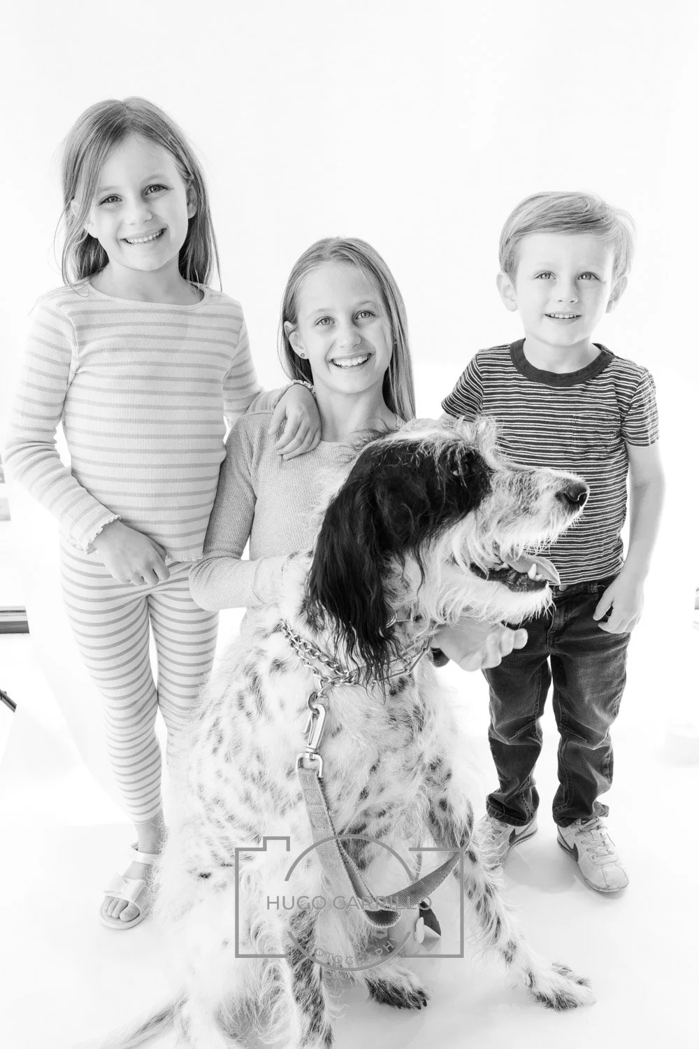 Three children, two girls and one boy, standing behind a large dog with a long fur coat, in a bright studio setting.