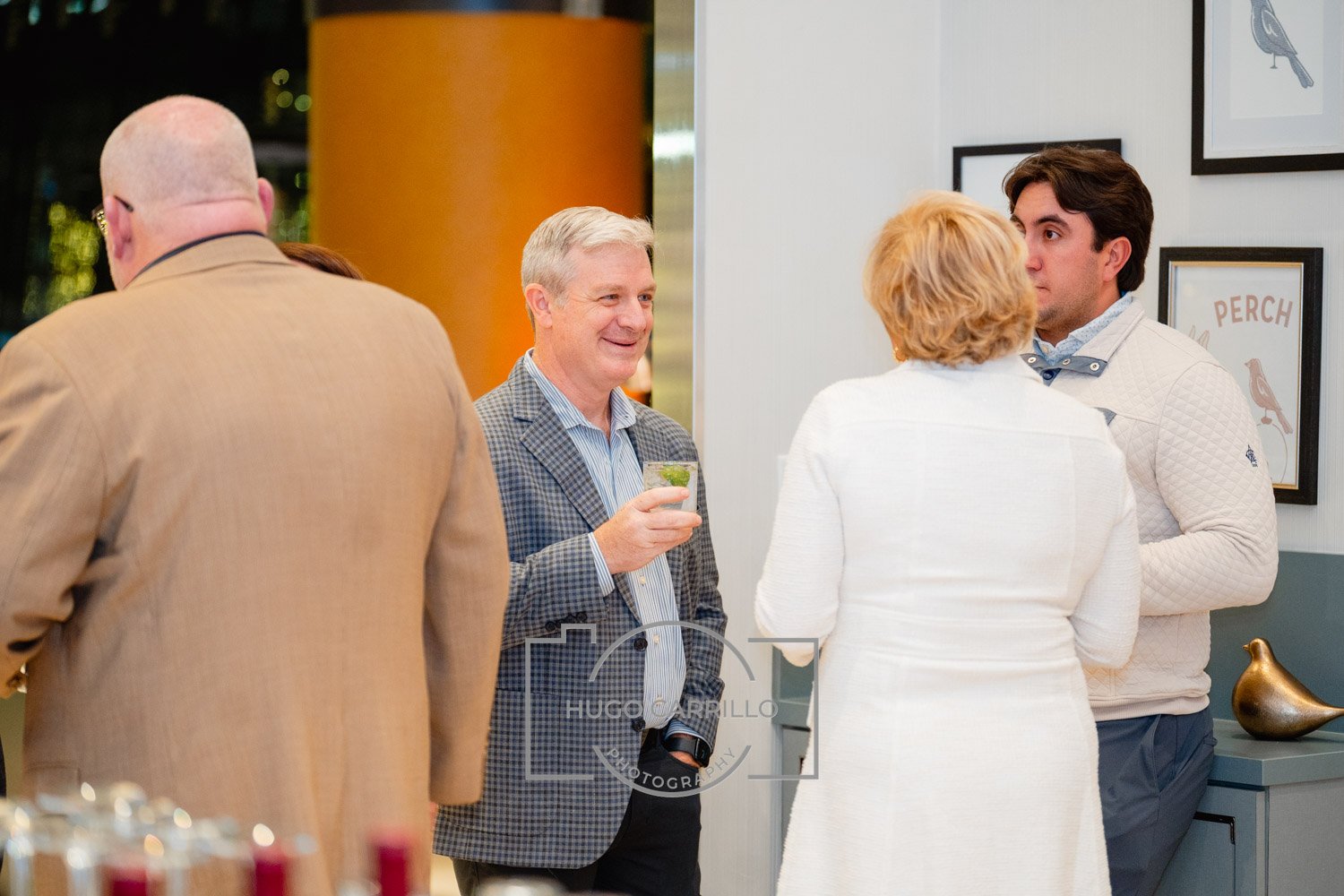 Group of five people socializing at an indoor event, with a man holding a drink smiling and talking to others