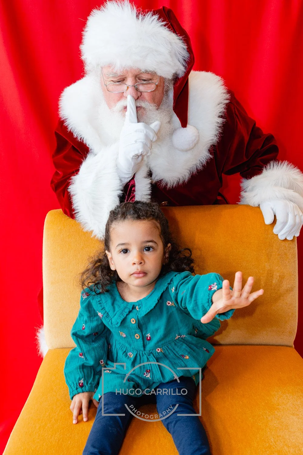 A girl with curly hair sitting on a yellow chair, reaching out with her hand. Behind her, Santa Claus with glasses and a white beard, wearing a red suit with white fur trim, is making a shushing gesture with his finger to his lips. The background is 