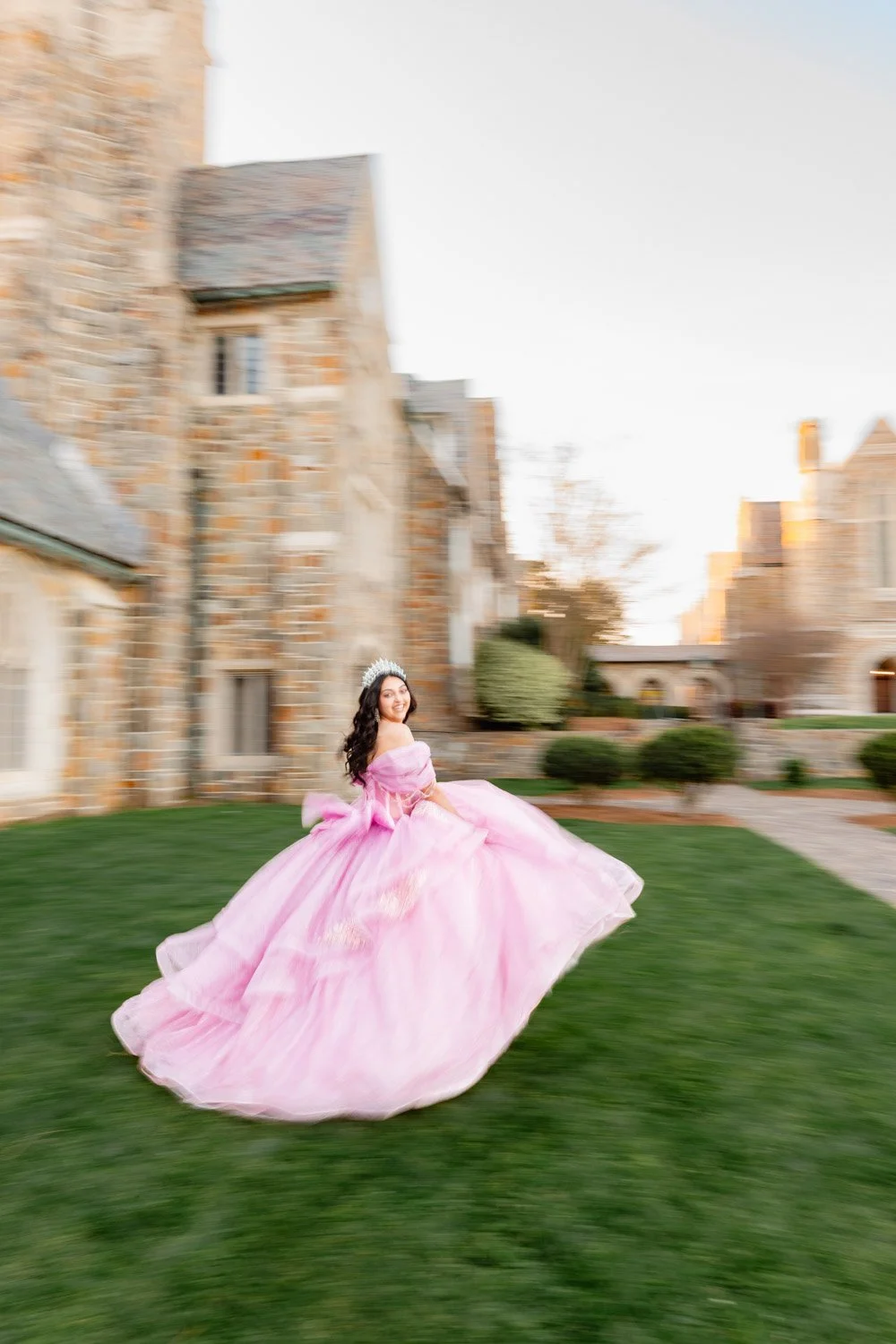 Young woman in a pink ball gown and tiara spinning on a lawn in front of a stone building.