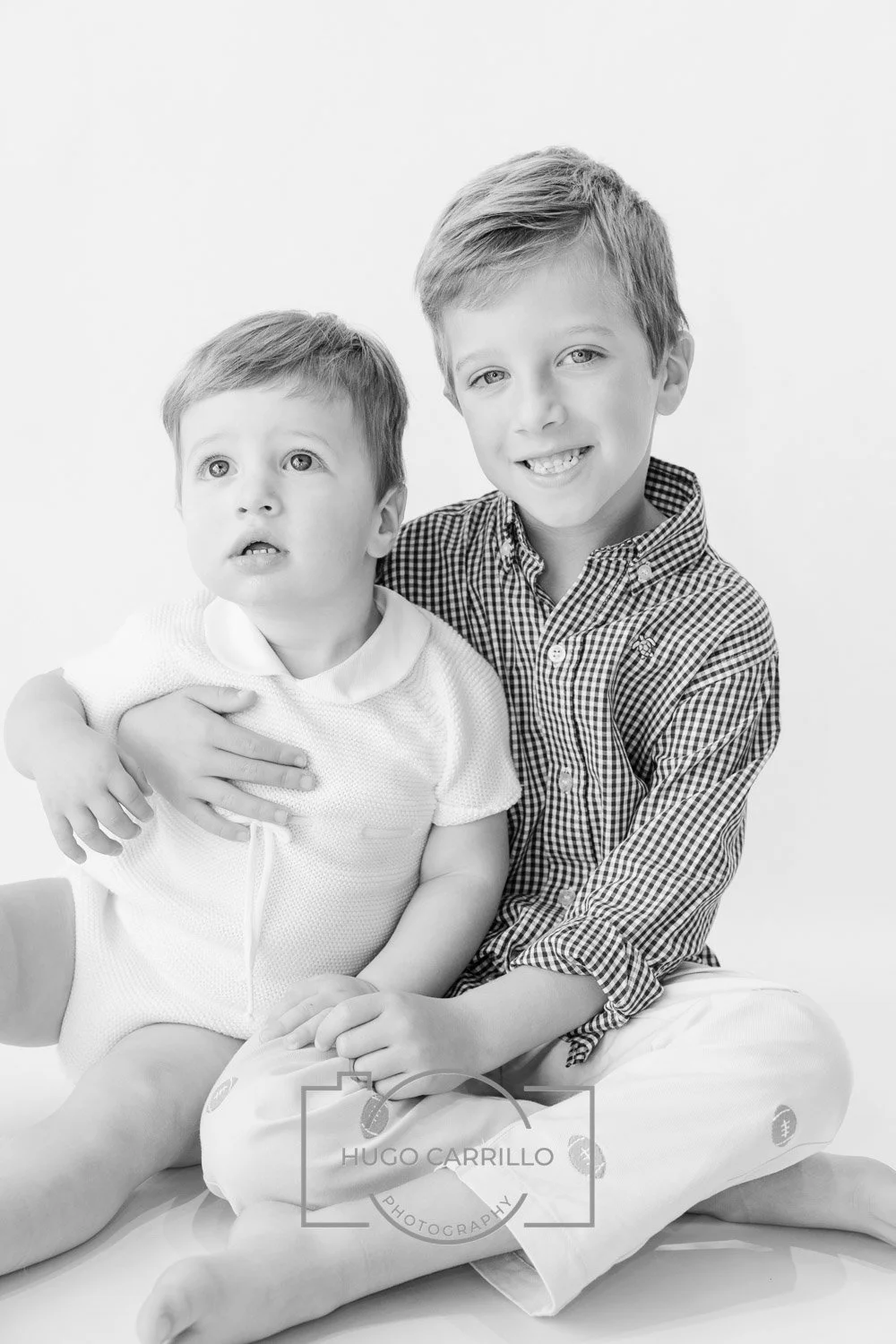 Two young boys, one older and one younger, sitting together on the floor. The older boy has short, light-colored hair and is smiling while holding the younger boy. The younger boy has similar light hair and looks curious. Both are dressed in casual c