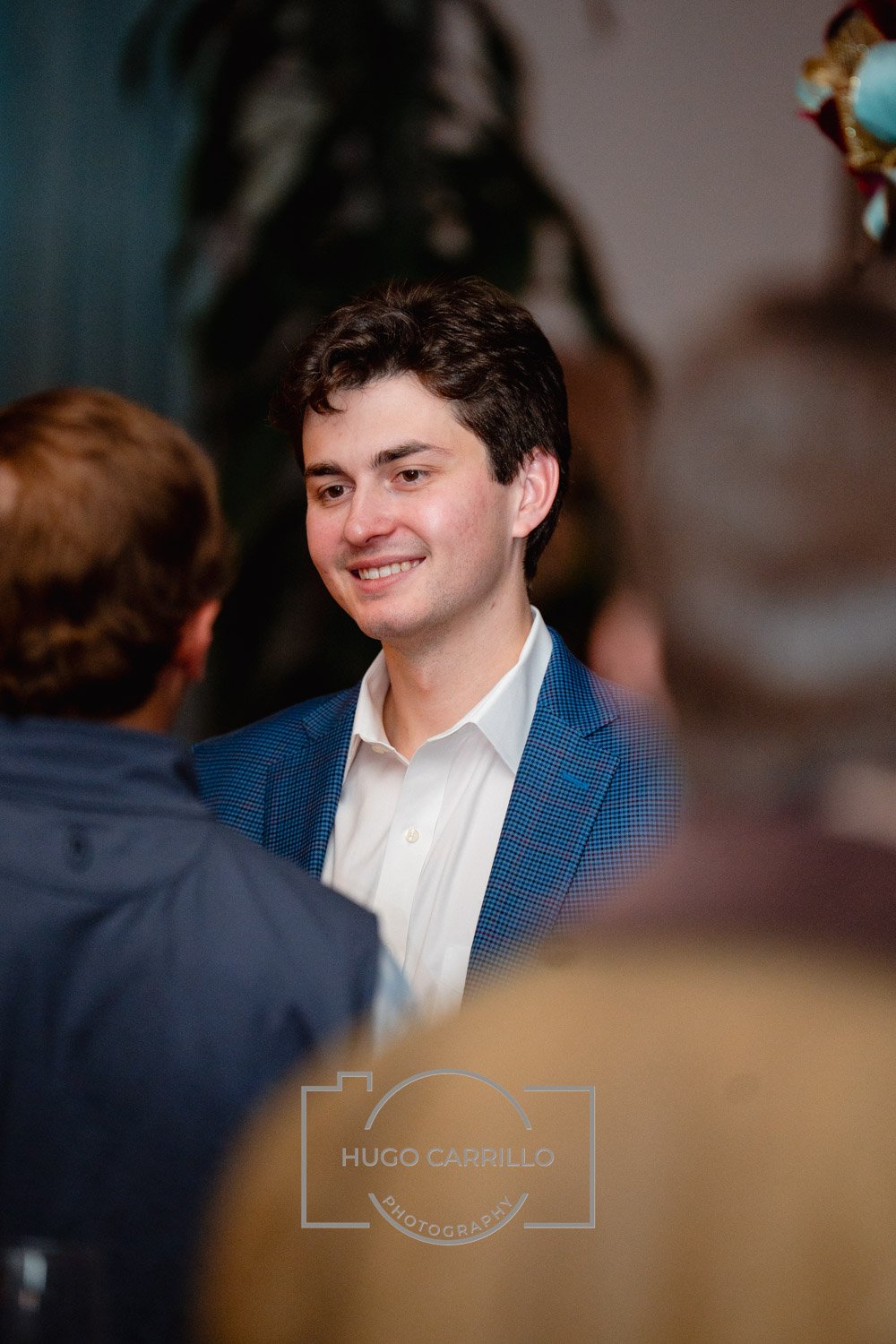 Young man with dark hair in a white shirt and blue blazer, smiling, engaged in conversation at an indoor social event, with blurred people around him.
