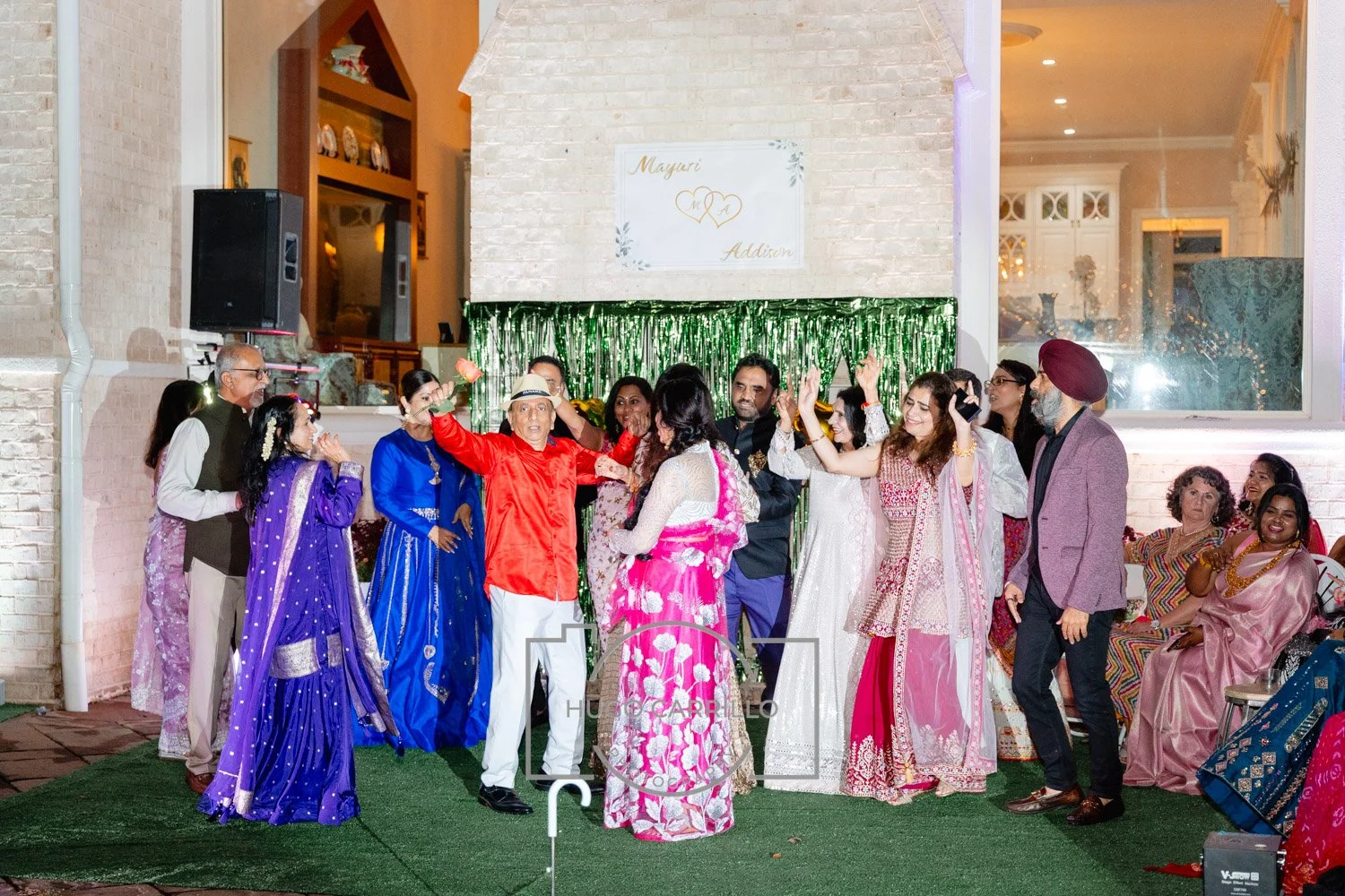 A diverse group of people celebrating at a wedding reception outdoors, with some dancing, wearing colorful traditional and modern attire, in front of a green tinsel backdrop and a sign with names and hearts.