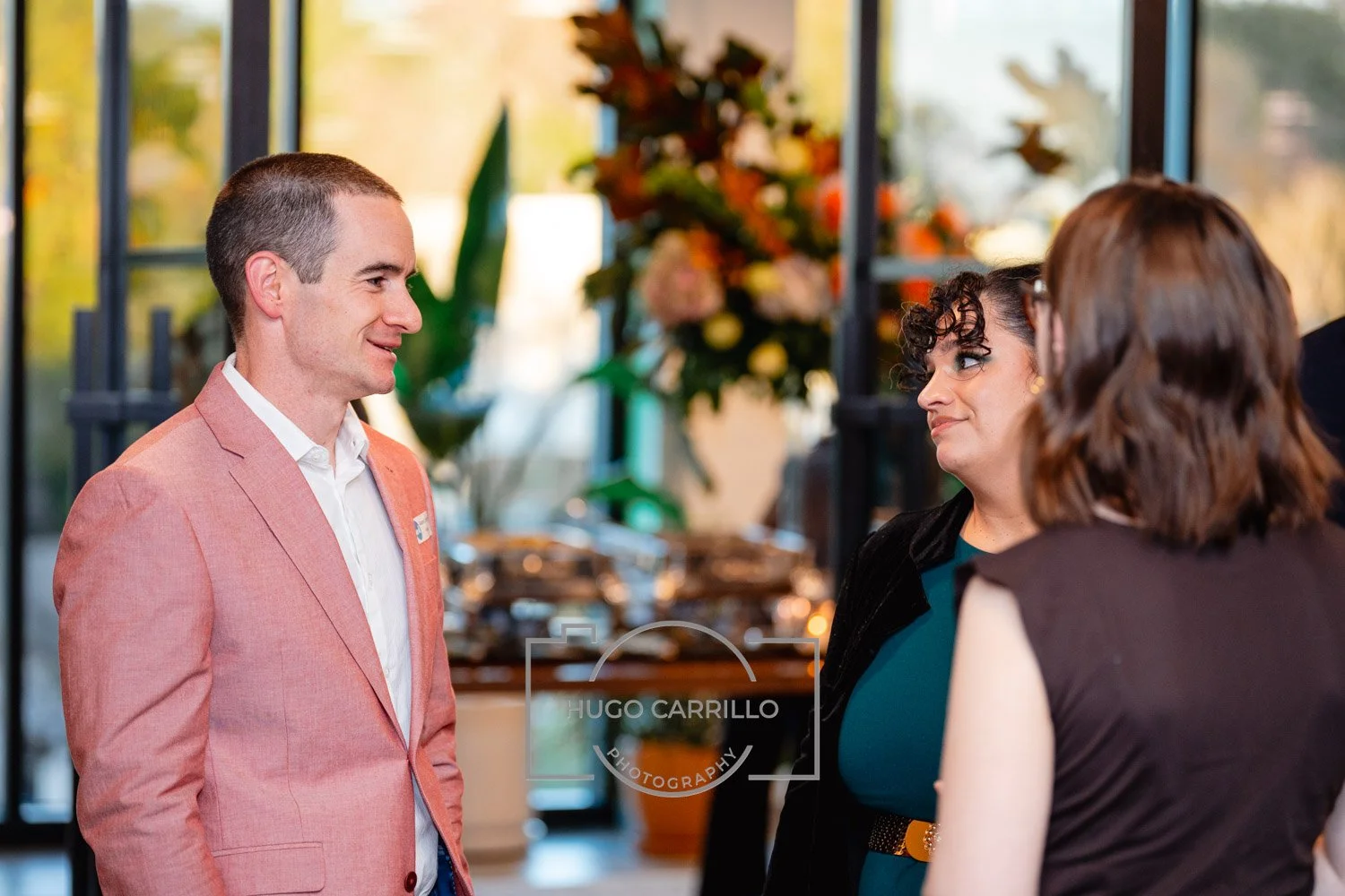A man in a light pink blazer smiling and talking to two women in a brightly lit indoor setting, with floral arrangements in the background.