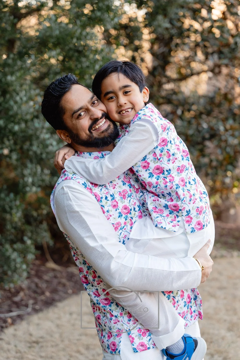 A man and a young boy, both with matching floral shirts, smiling and hugging outdoors with trees and sunset in the background.