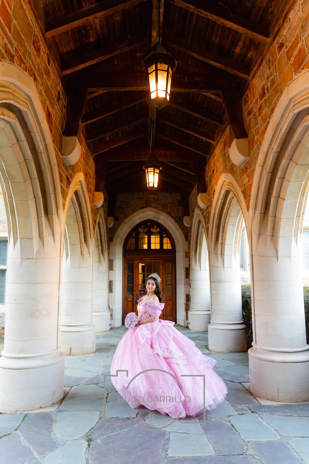 A young woman in a pink quinceañera dress with a crown and tiara, holding a bouquet, standing under a stone archway with hanging lanterns.