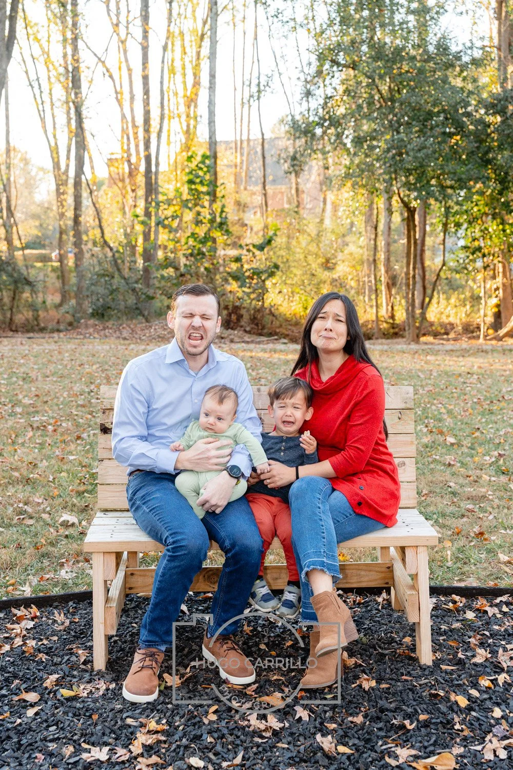 A family of four sitting on a wooden bench outdoors during fall, with trees and fallen leaves in the background. The father is holding a baby, and the mother is sitting next to a young boy, all with emotional or distressed expressions.