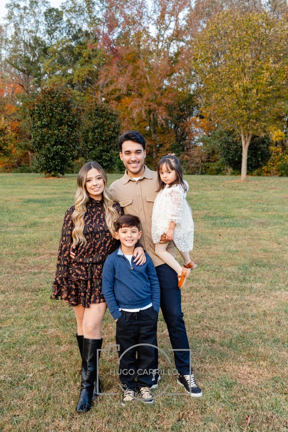A family of four standing on grass in a park with autumn trees in the background. The father holds a young girl, the mother stands next to them, and a young boy stands in front. All are smiling and dressed in casual fall clothing.