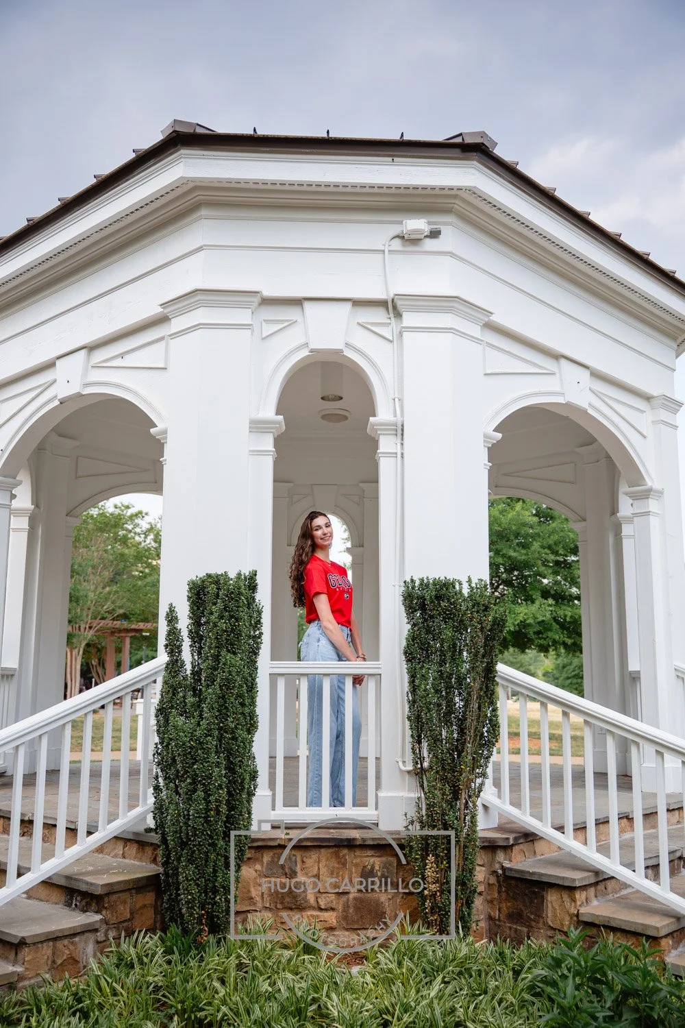 Woman standing on a small white porch with railing, in front of a white gazebo, wearing a red t-shirt and blue jeans, smiling.