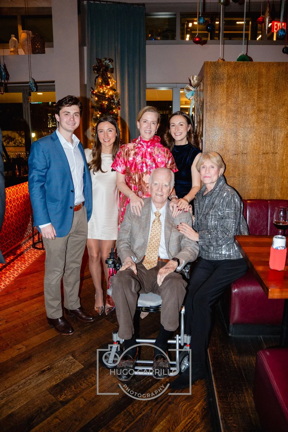 Group of six people, including an elderly man in a wheelchair, posing together at a family gathering or celebration in a restaurant, with holiday decorations in the background.