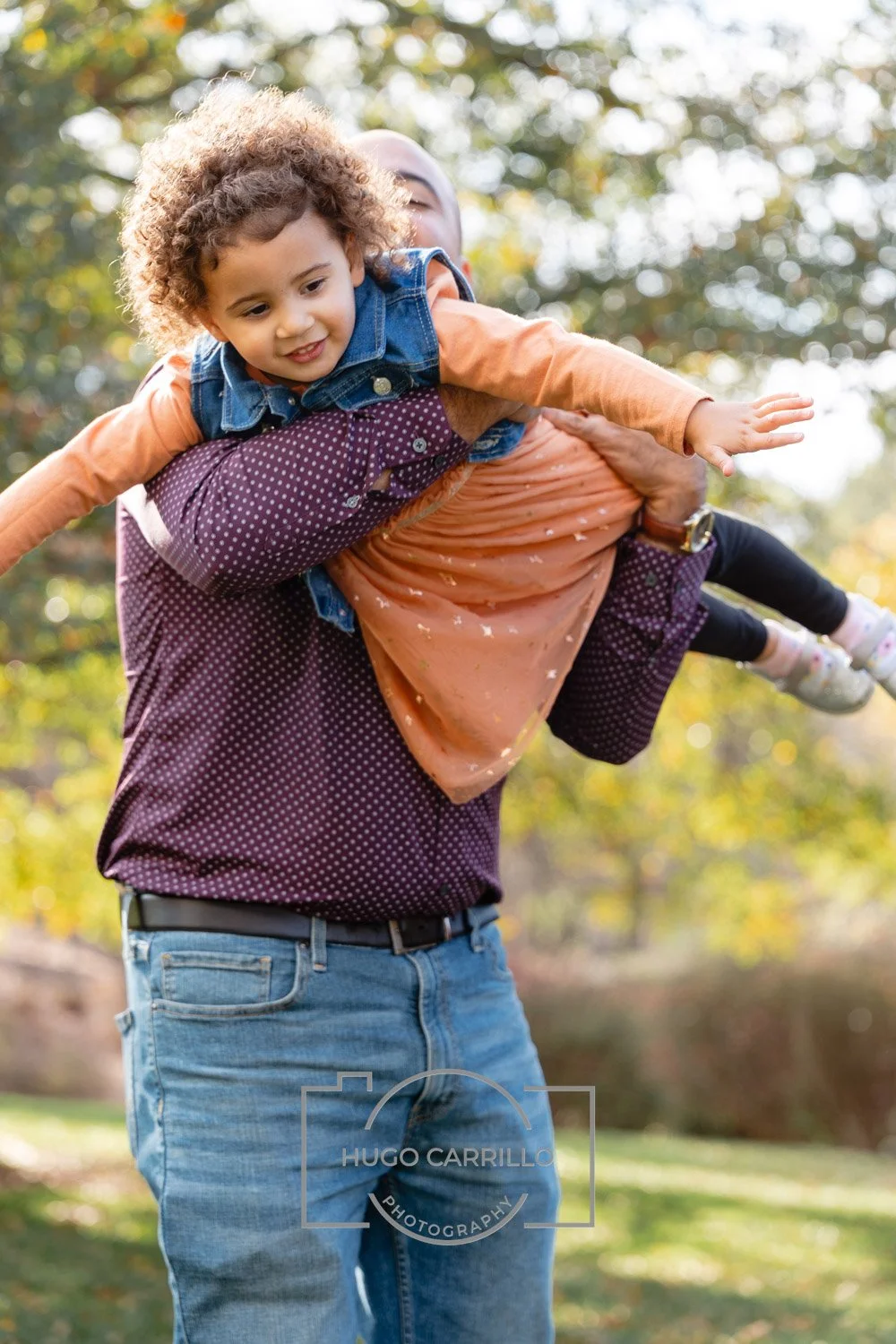 A person holding a young child on their shoulder outdoors with trees and sunlight in the background.